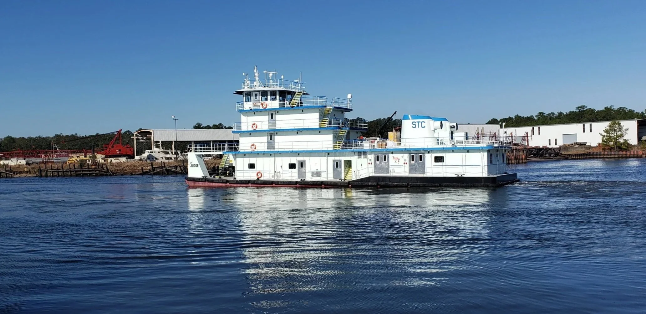 A white and blue riverboat with multiple decks sailing on calm water near a dock and industrial buildings in the background.