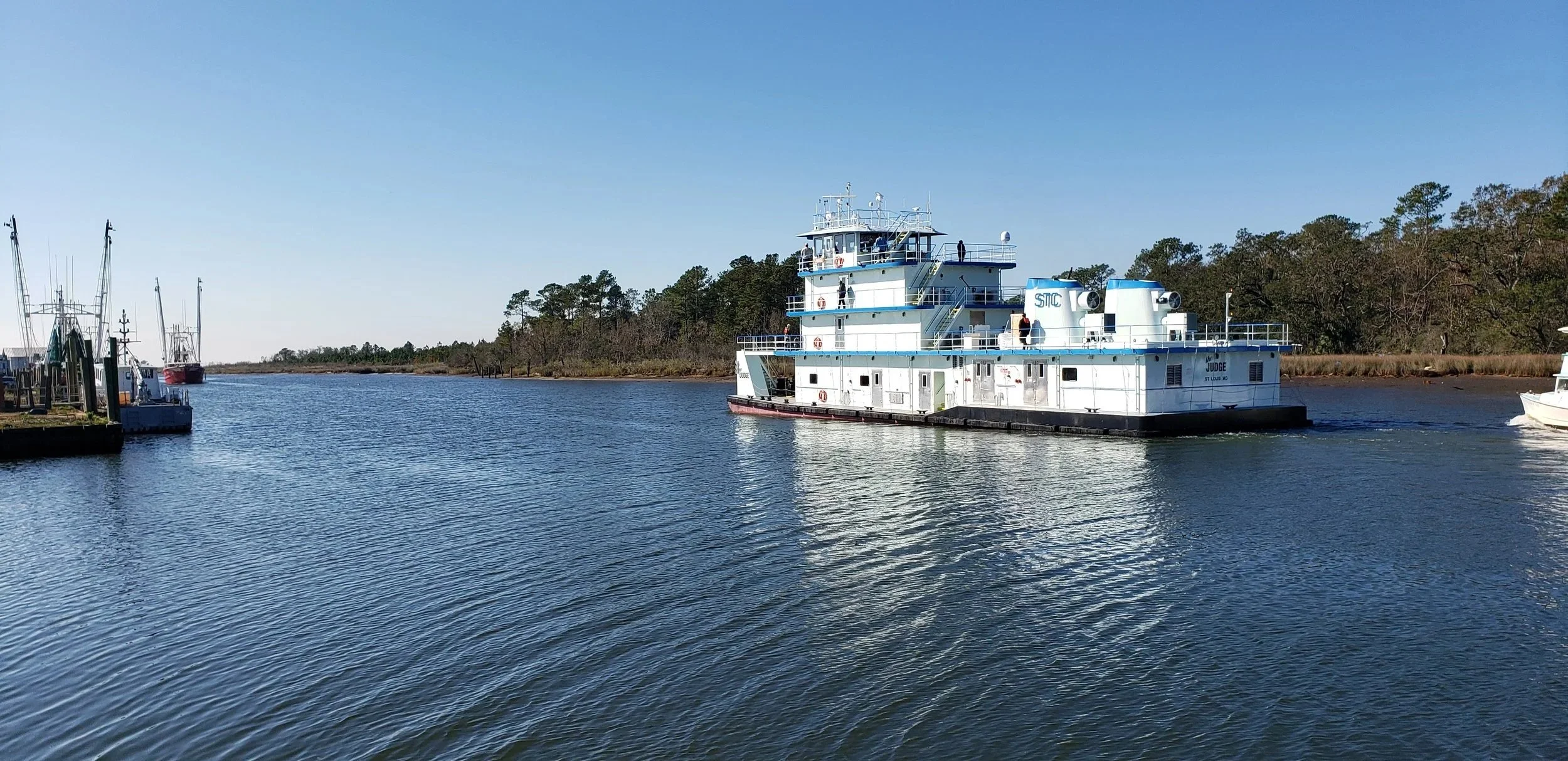A large white boat named 'Judge' sailing on a calm body of water, with a shoreline and trees in the background under a clear blue sky.