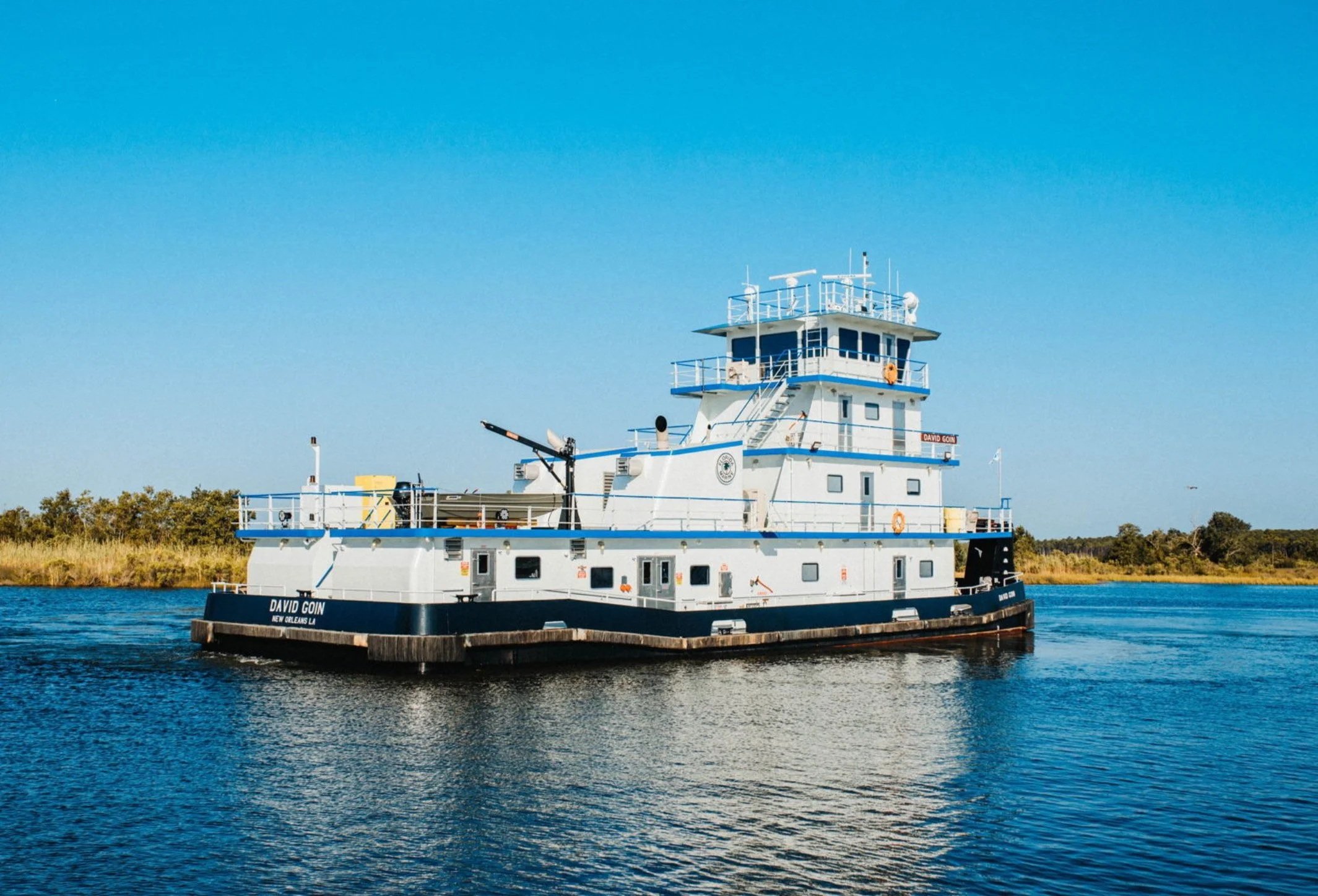 A white and black tugboat named David Goin on a body of water with a grassy shoreline and trees in the background under a clear blue sky.