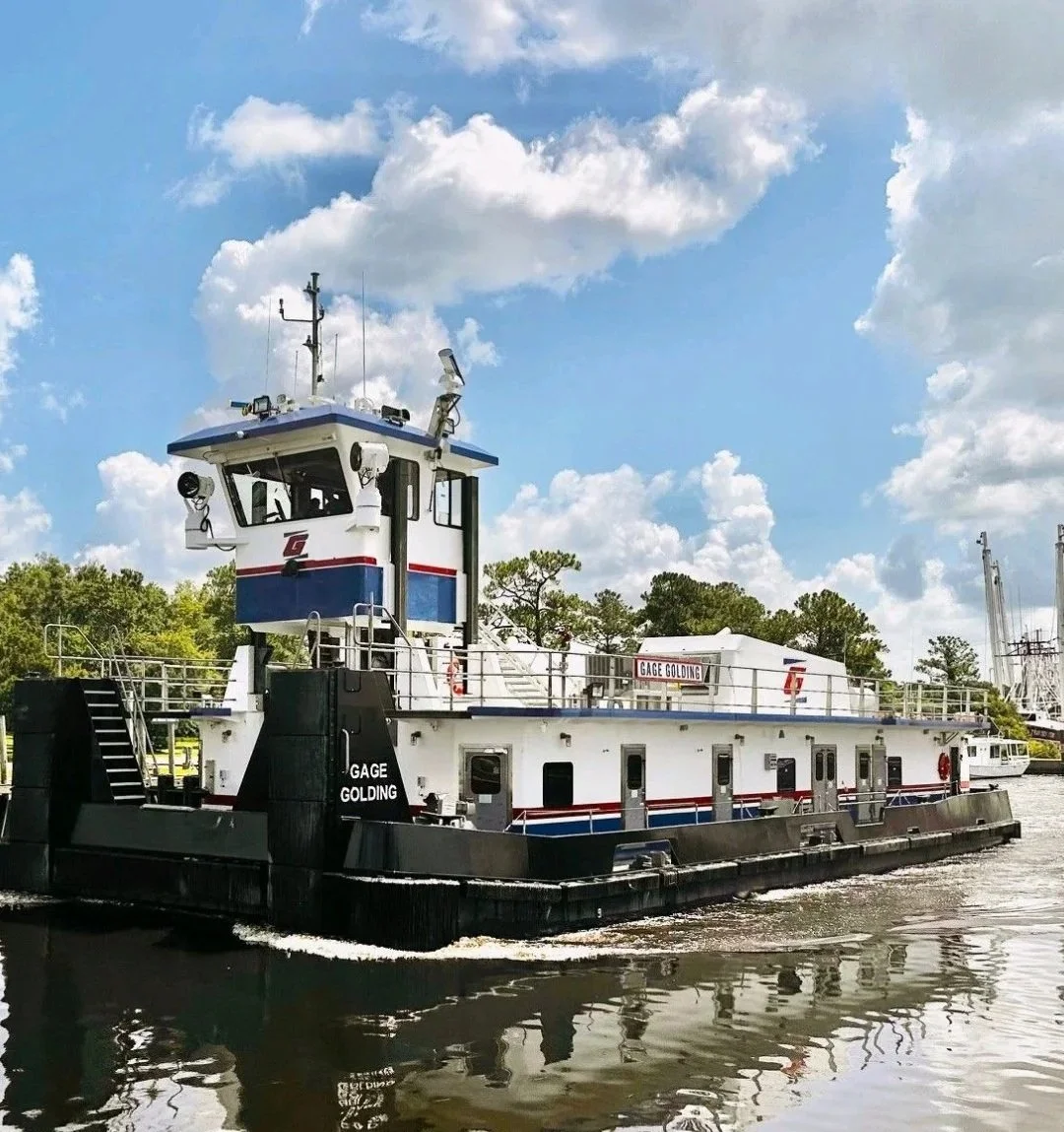 A towboat named Gage Golding on a river with blue sky and clouds above, surrounded by green trees in the background.