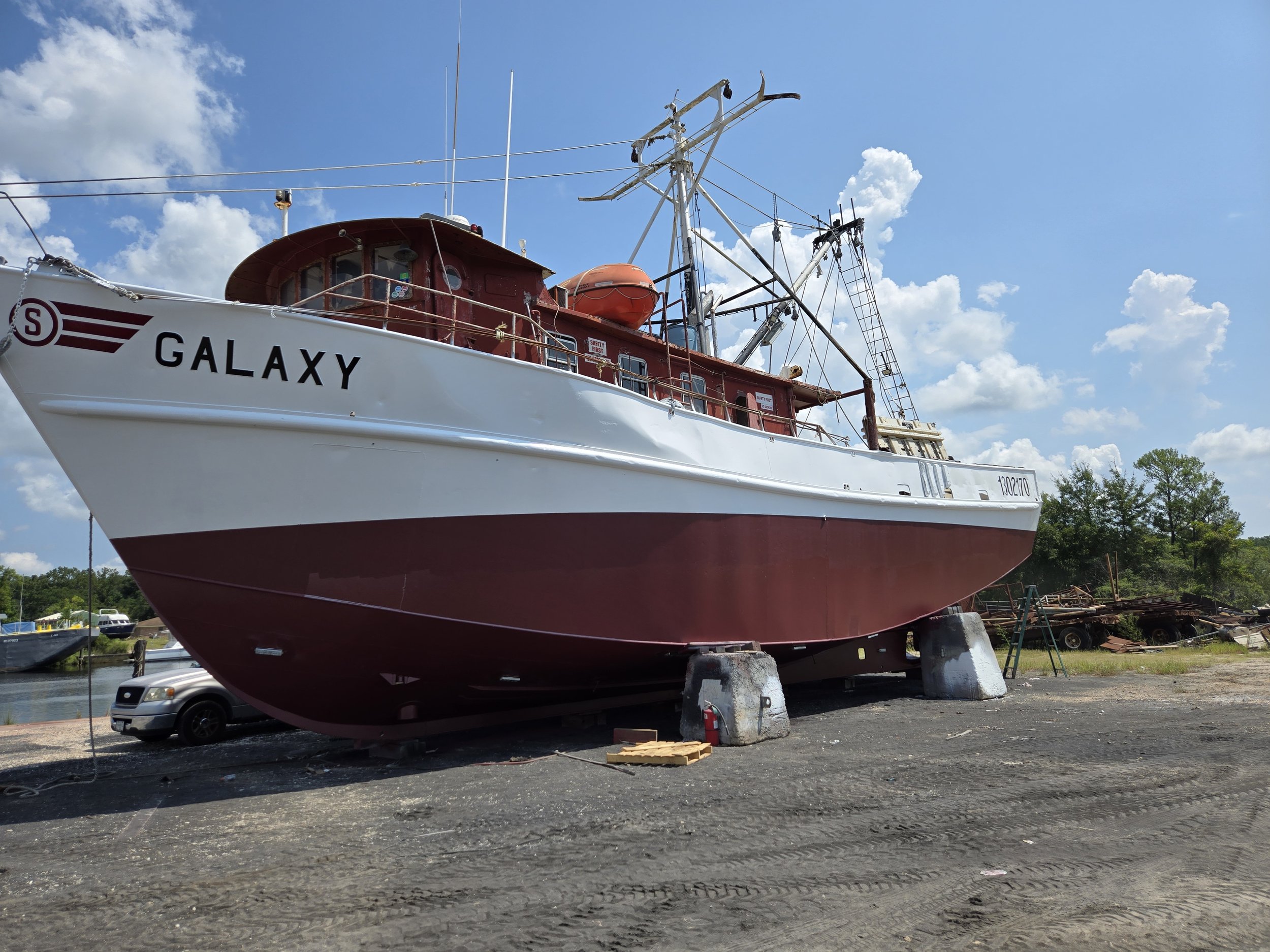 A large red and white boat named 'Galaxy' on a dry dock, with a blue sky and clouds above, and some trees and miscellaneous items in the background.