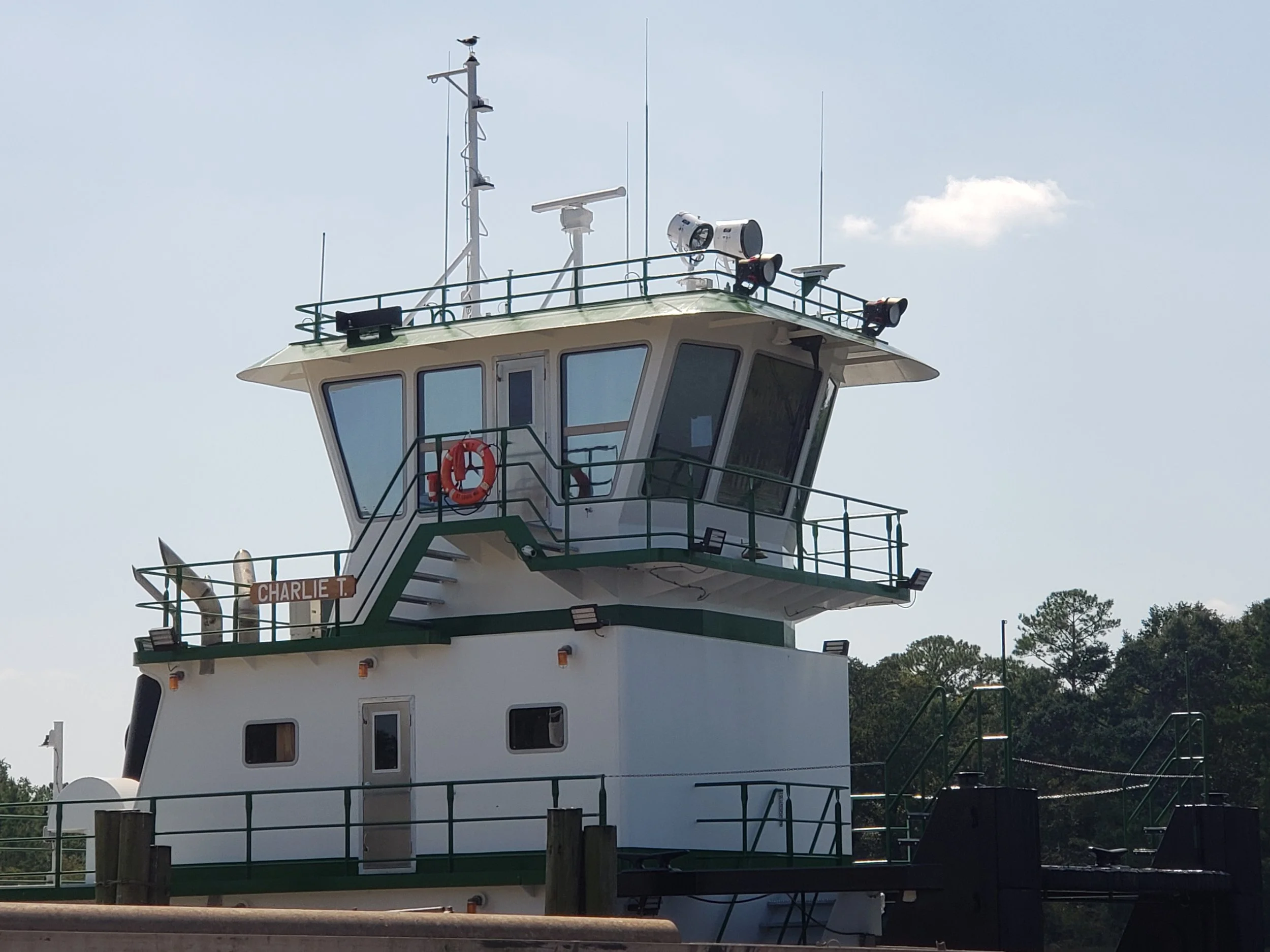 A white boat control tower with large windows, green railings, and multiple antennas, set against a partly cloudy sky with trees in the background.