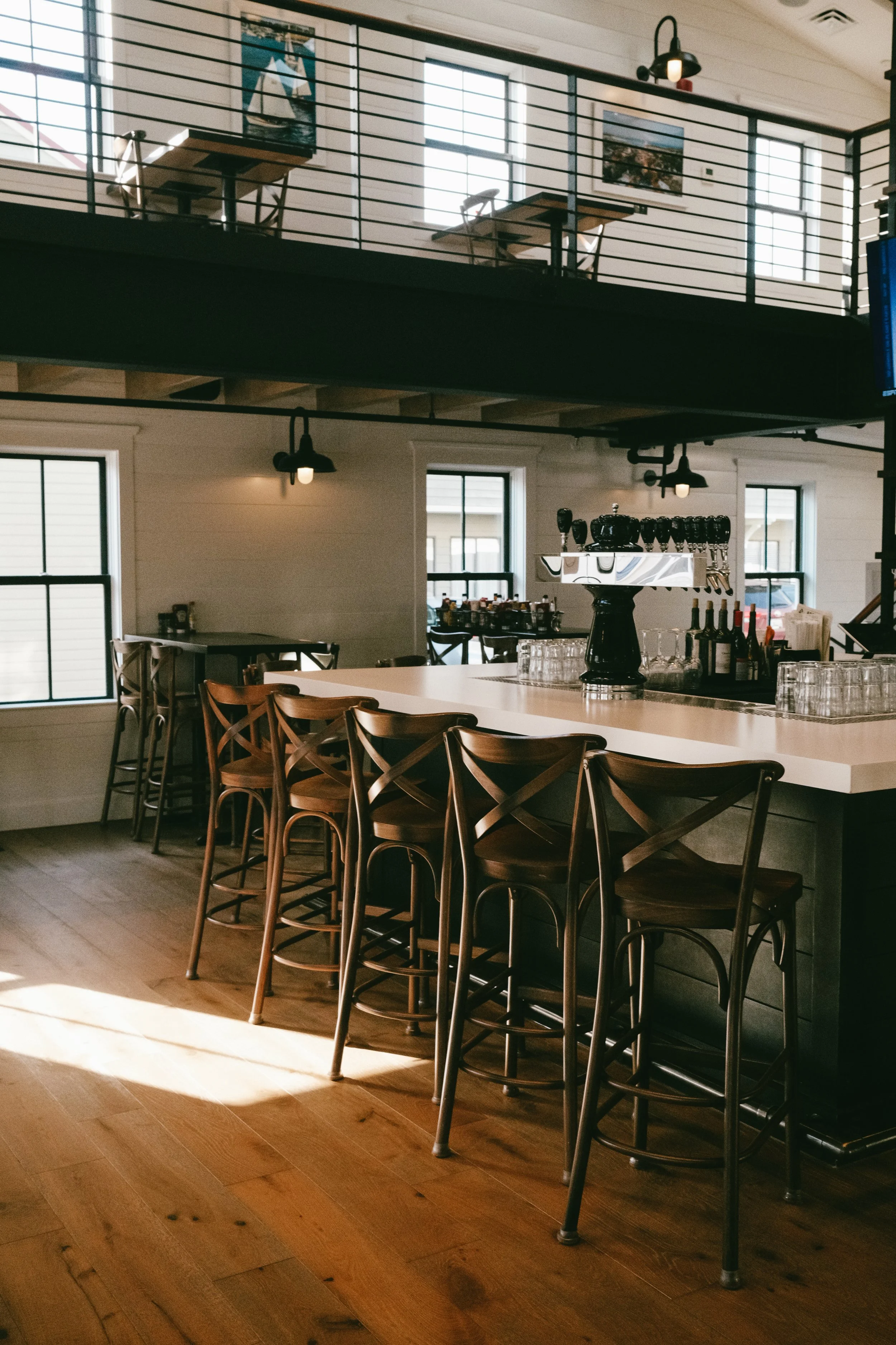 Interior of Adam's on Fourth in Eastport restaurant with a white counter and wooden chairs. The counter has a coffee machine and glassware, with large windows in the background and black lighting fixtures hanging from the ceiling.