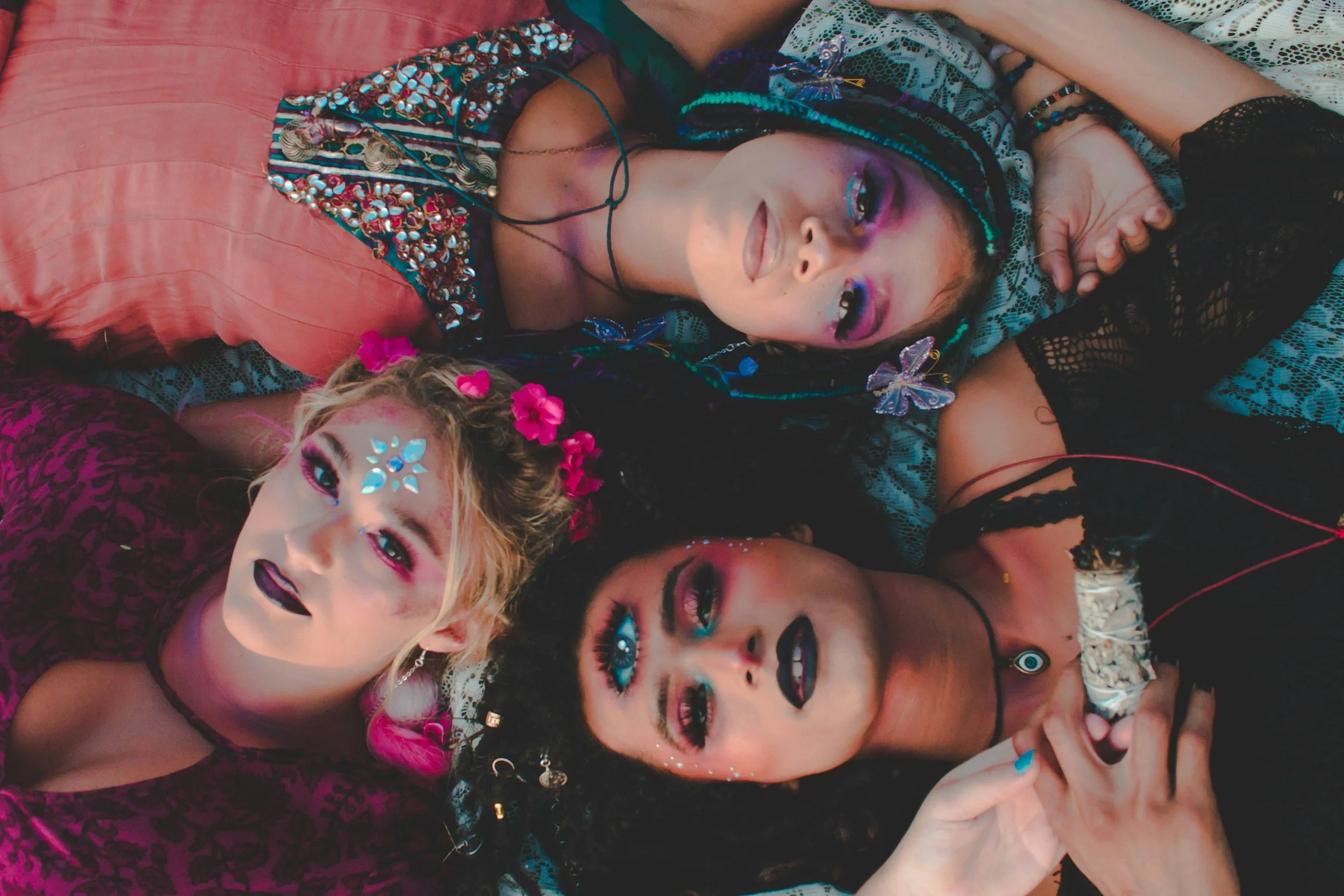 A group of young women lying close together, dressed in colorful and elaborate outfits with bright makeup and accessories, posing for a selfie.