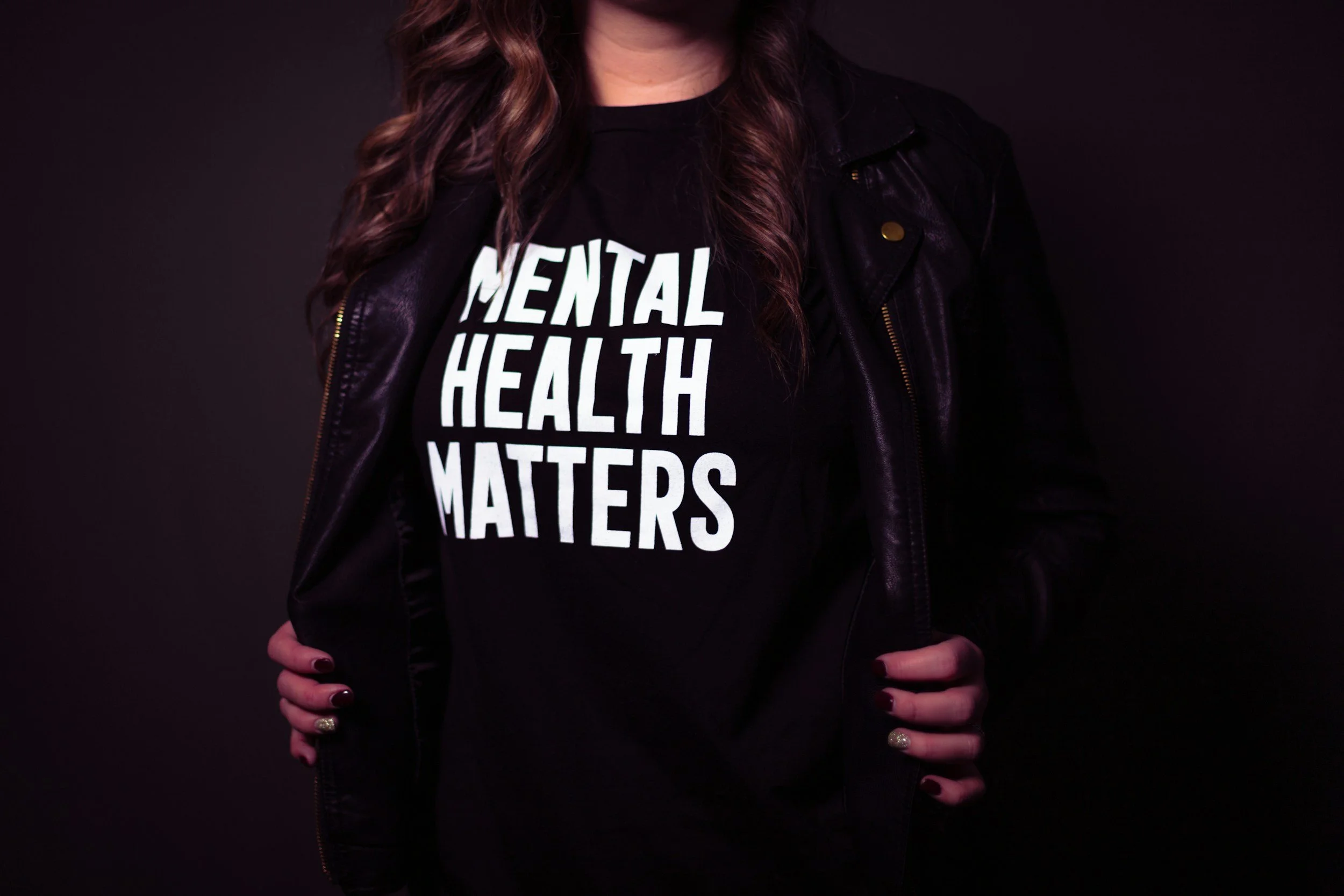 Person wearing a black t-shirt with white text that reads 'MENTAL HEALTH MATTERS' and a black leather jacket, with long wavy hair and painted nails.
