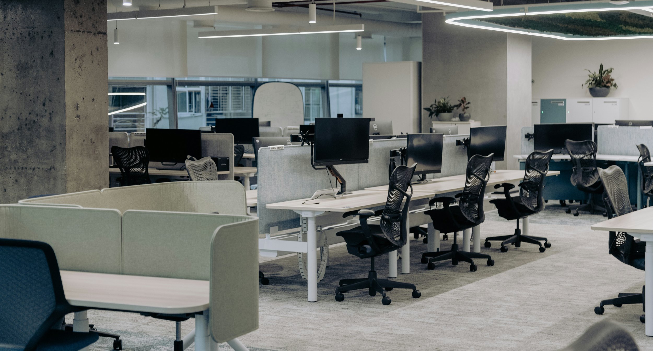 An empty modern office with multiple workstations, black ergonomic chairs, and desktop monitors. The space features a mix of cubicles, a concrete pillar, and large windows, with plants and storage cabinets visible.