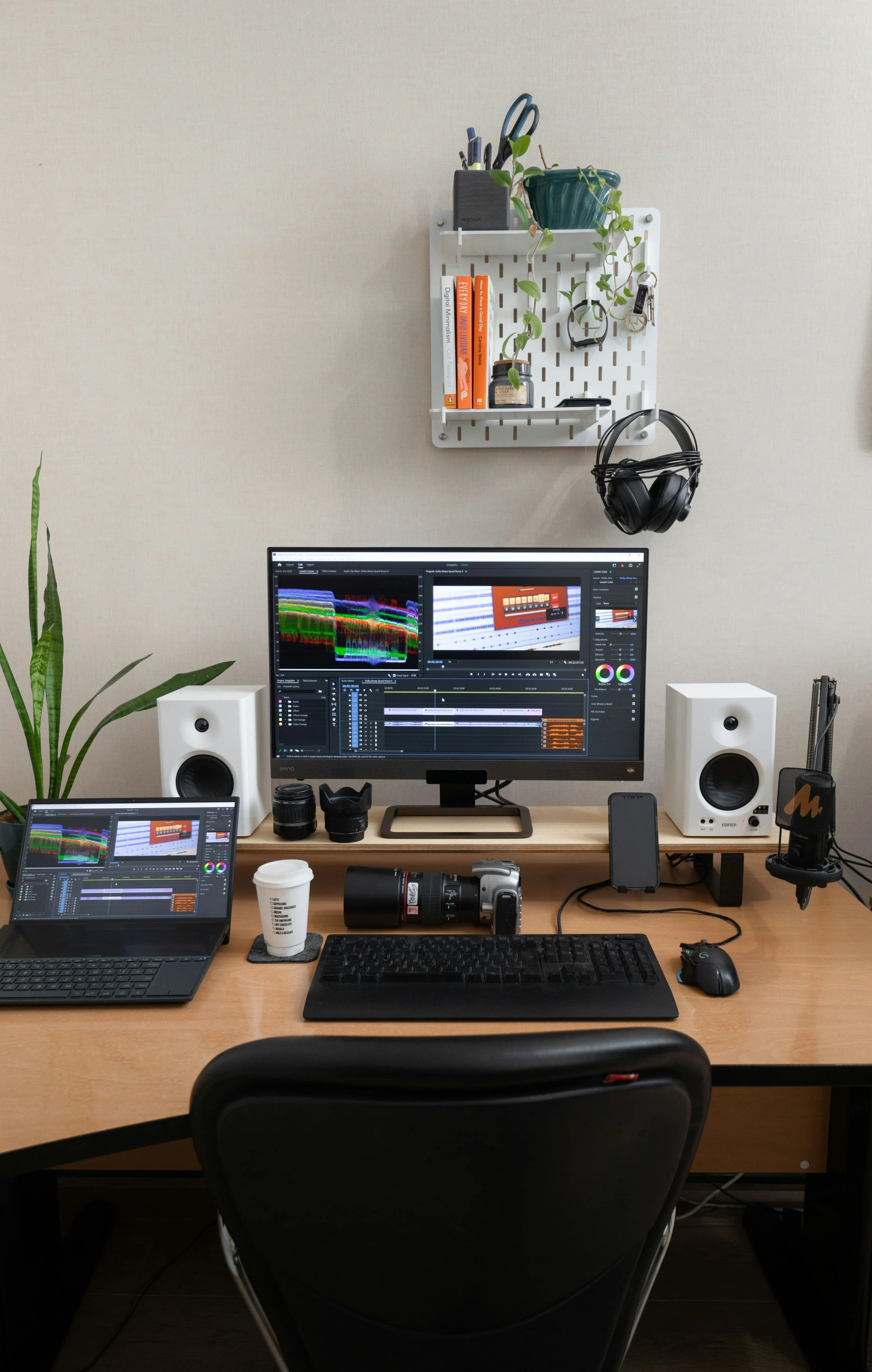 A workspace with dual computer monitors displaying audio editing software, a laptop, camera lenses, a coffee cup, a camera, a keyboard, mouse, speakers, headphones, a smartphone, a microphone, and a green plant on the desk. A wall-mounted white pegboard holds books, pens, a plant, and keys.