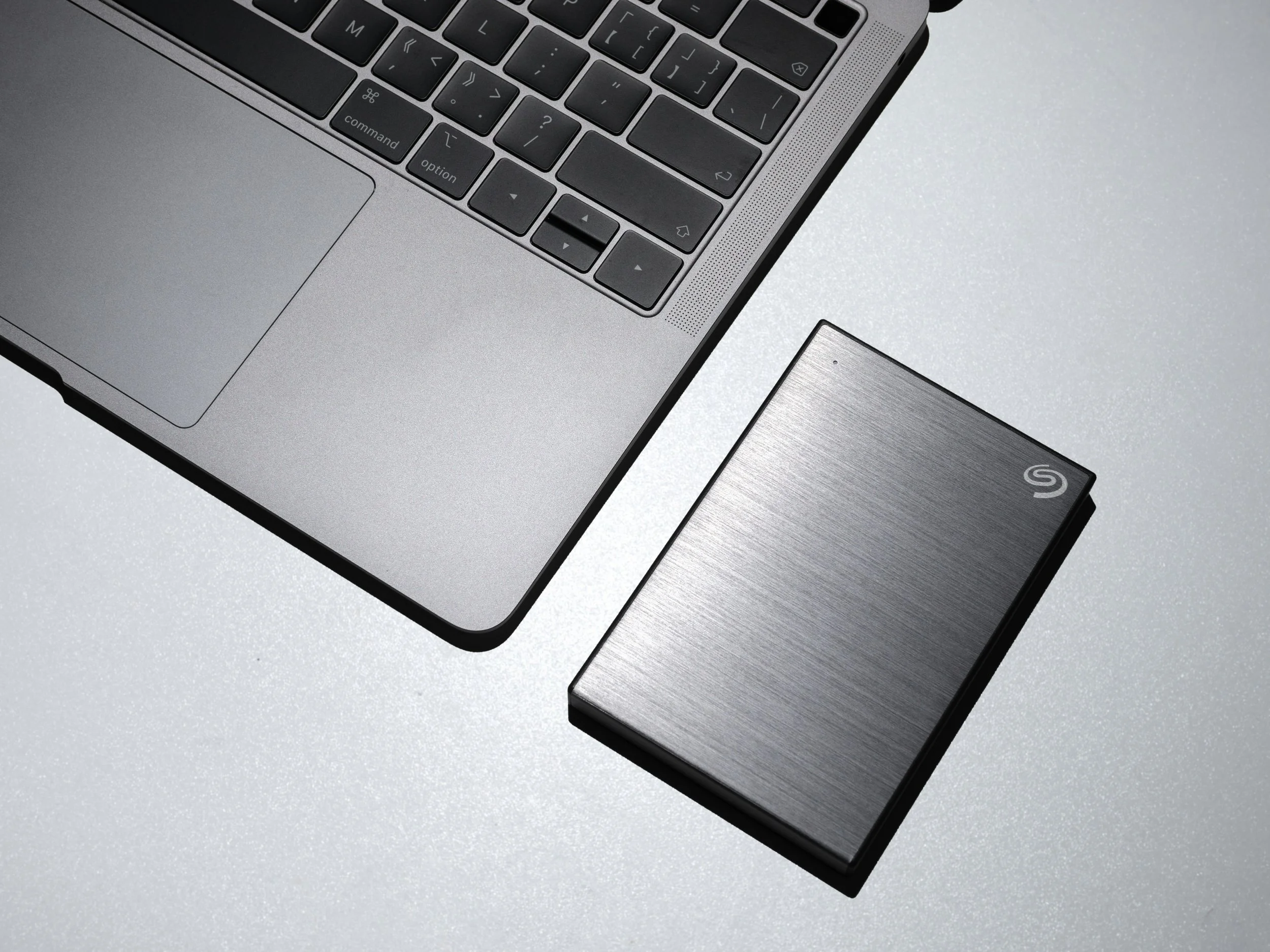 Close-up of a silver laptop keyboard and a portable external solid-state drive (SSD) on a light-colored surface.