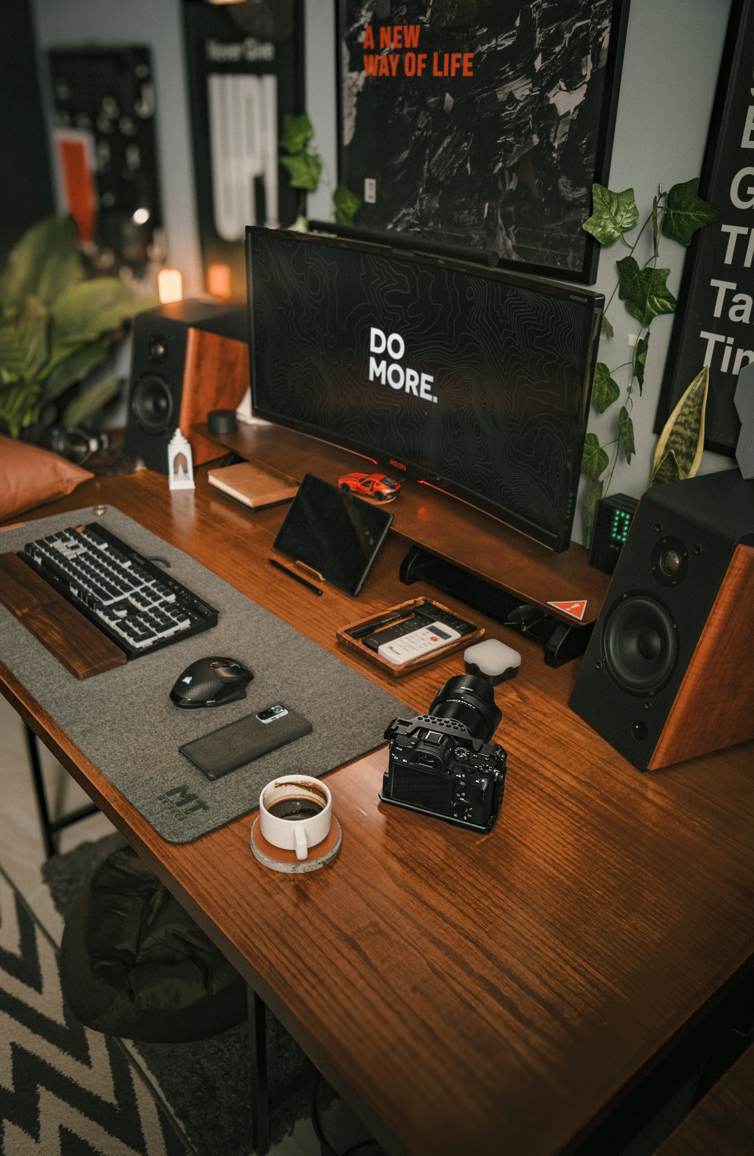 A wooden desk with a computer monitor displaying the words 'DO MORE.' In front of the monitor are a keyboard, a mouse, a smartphone, a camera, and a cup of coffee. There are speakers on both sides of the monitor, and art posters on the wall behind.