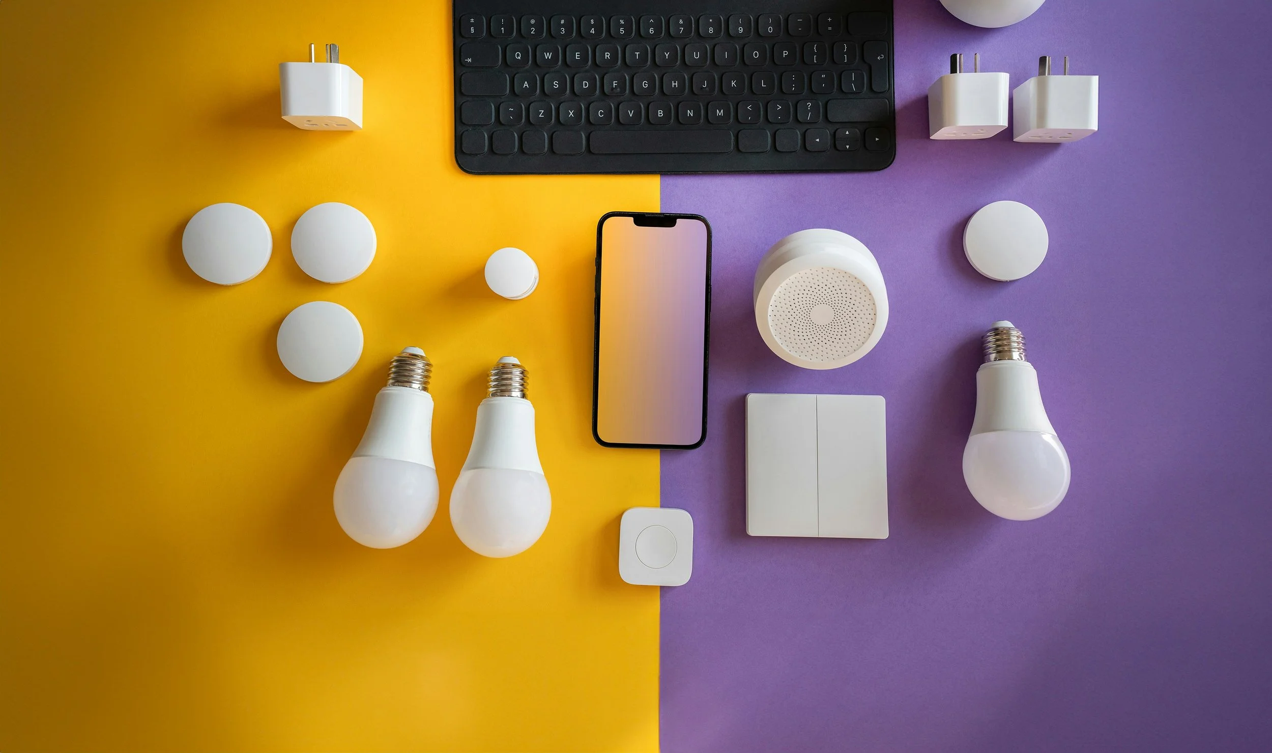 Top-down view of various electronic devices and light bulbs on a split yellow and purple background. Includes a black keyboard, a smartphone, two white smart plugs, several white light bulbs, and power adapters.