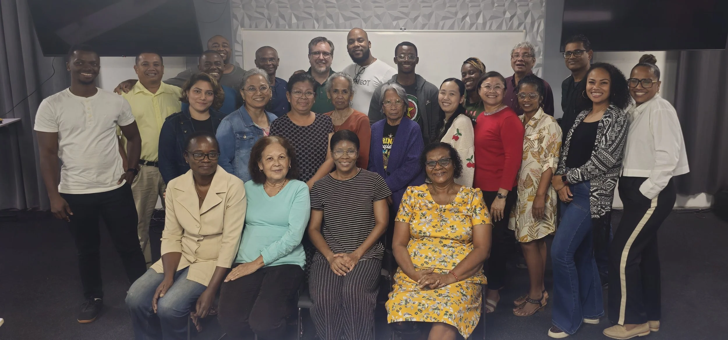 Group of diverse men and women of various ages posing together in an indoor setting, some seated and some standing, with a whiteboard and large screen in the background.