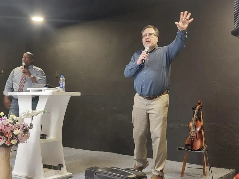 Two men speaking at a podium, one with a microphone and the other with a hand raised, on a stage with a violin on a stand nearby and a flower arrangement in the foreground.