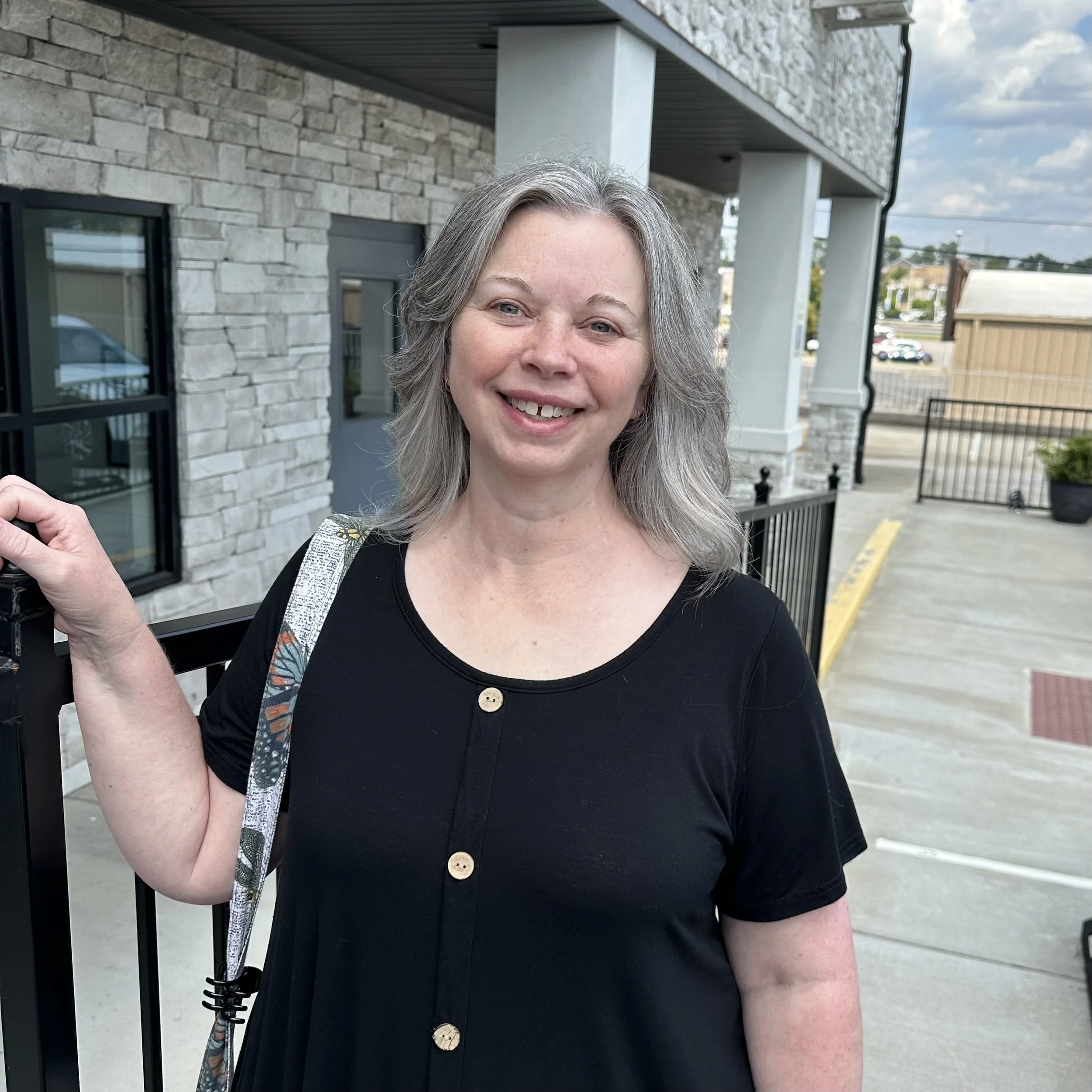 A smiling woman with grey hair standing outdoors near a metal gate, wearing a black dress with wooden buttons and carrying a patterned bag on her shoulder.