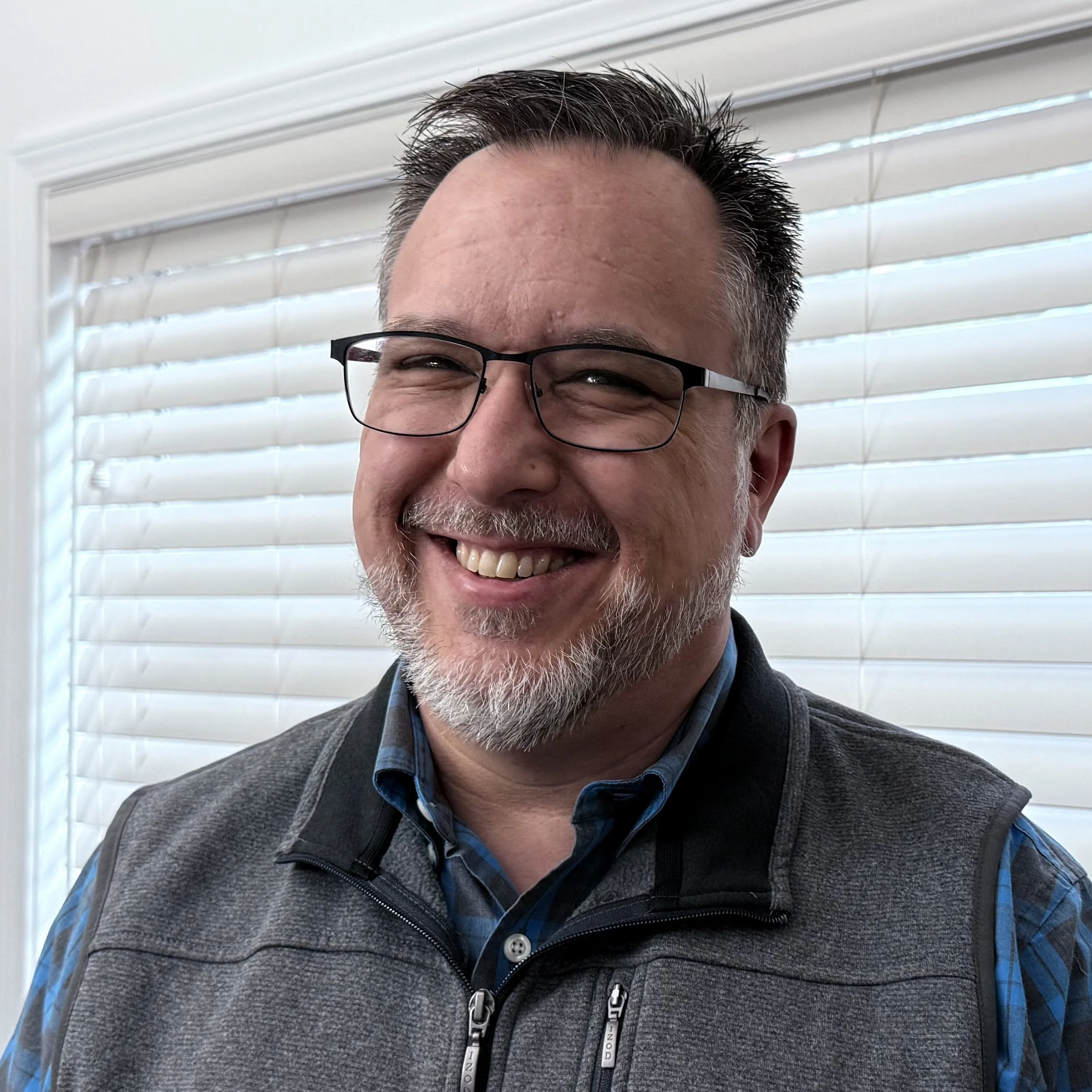 A smiling man with glasses and a beard standing in front of white window blinds.