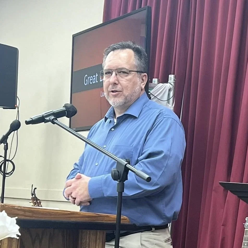 A man with glasses and a beard stands at a podium, speaking into a microphone. He is wearing a blue dress shirt and standing in front of a large television screen and red curtains.