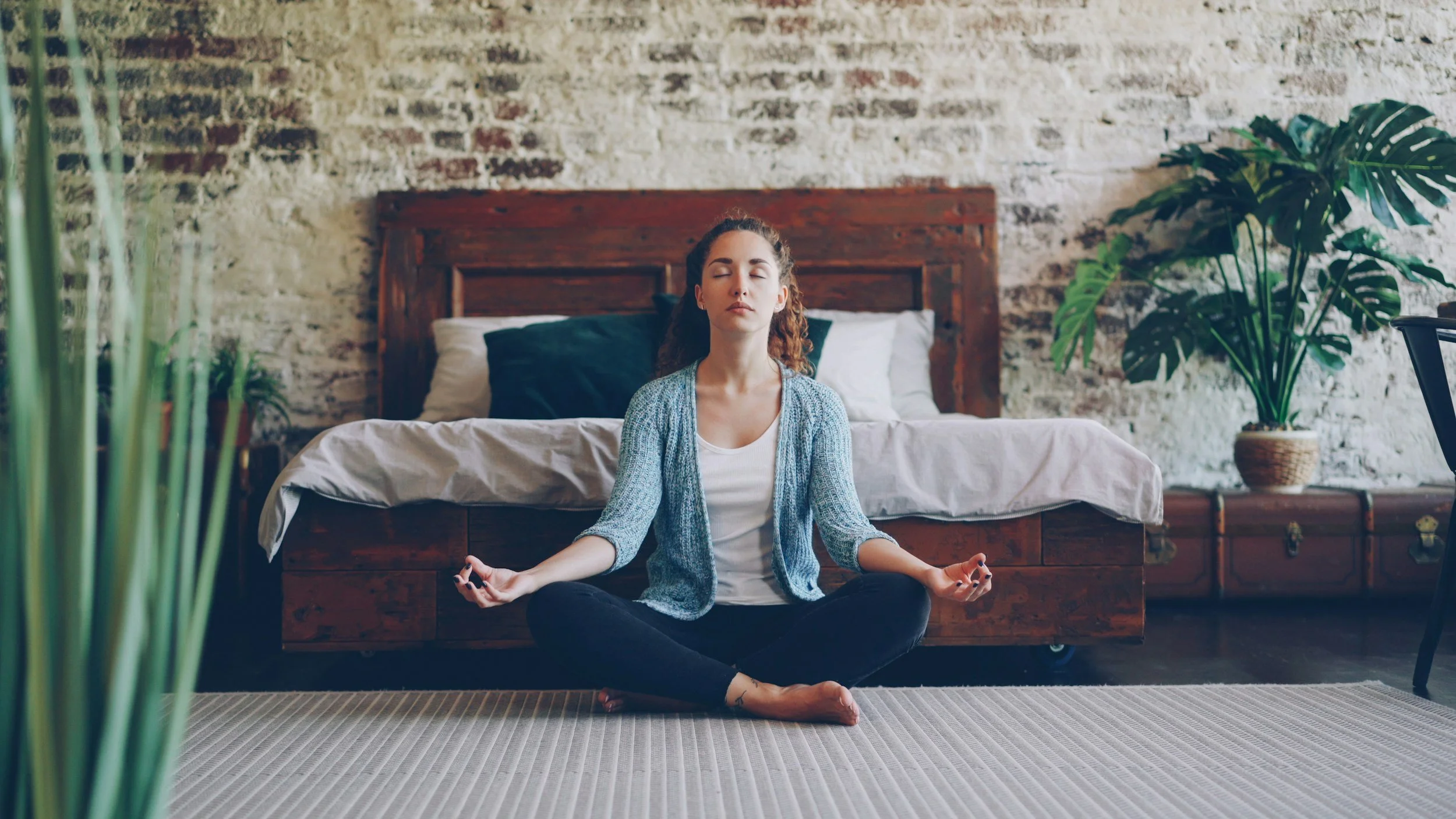 A woman sitting cross-legged on the floor in a bedroom, meditating with eyes closed, in front of a bed with a wooden headboard and large potted plants on either side.