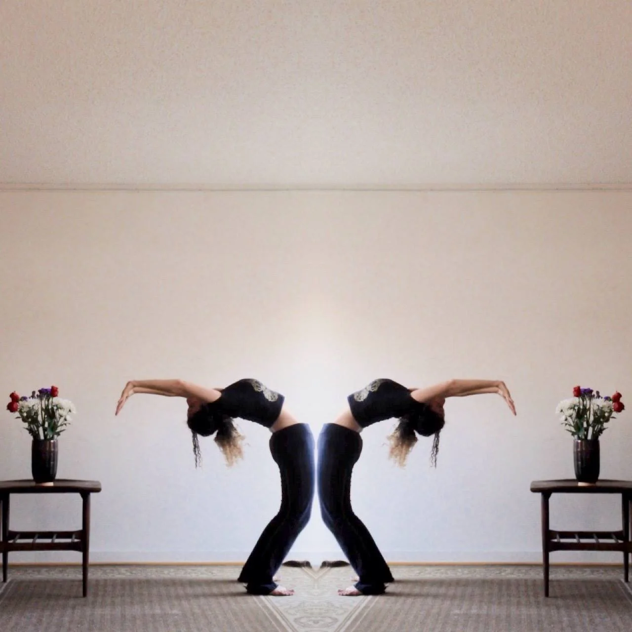 A mirrored image of two women in yoga poses, bent at the waist with arms extended downward, facing each other in a room with a beige wall, small wooden tables with vases of flowers on either side.