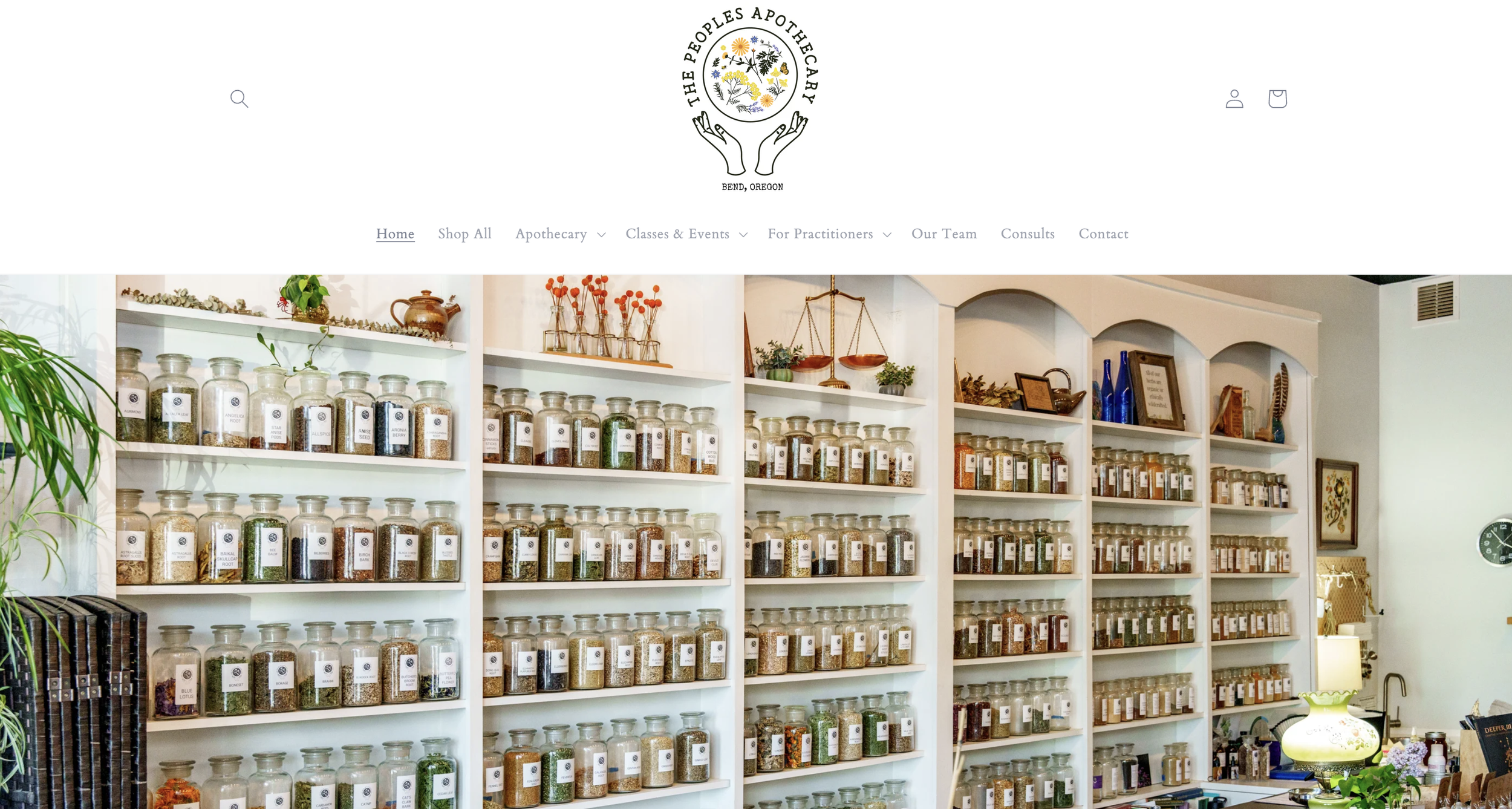 Interior view of a herbal apothecary shop with white shelves filled with glass jars containing herbs and spices, decorative plants, and framed artwork.