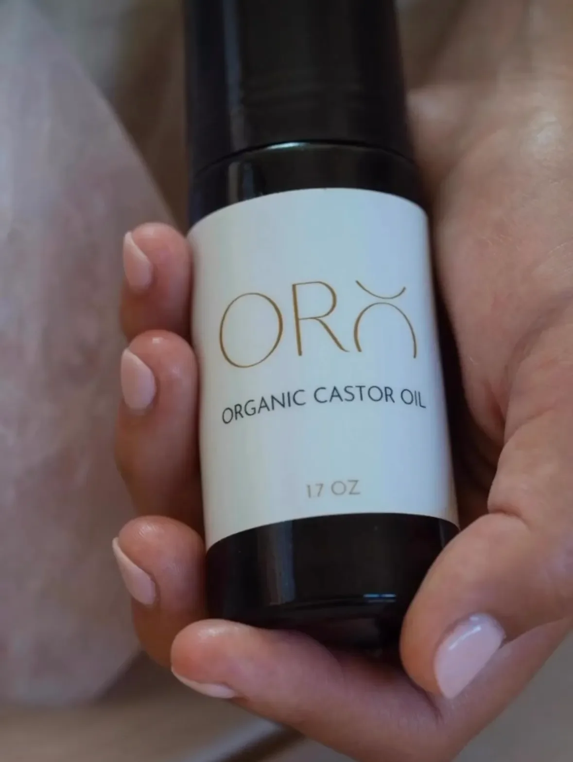 Hand holding a bottle of organic castor oil labeled 'ORA' with a black cap.