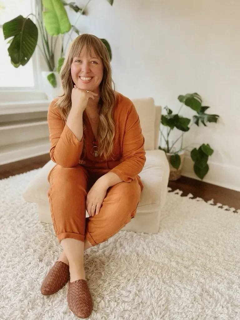 A woman with long blonde hair smiling, sitting on a beige armchair in a bright room with large green plants and white walls.