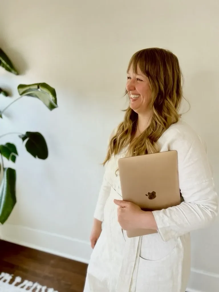 A woman with long, wavy hair smiling and holding a closed laptop while standing indoors near a large plant.