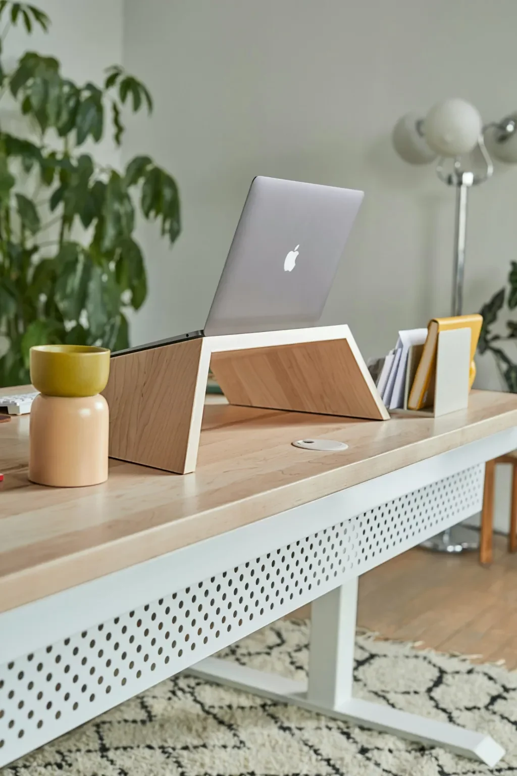 A workspace with a wooden desk holding an Apple laptop on a wooden stand, a yellow and beige ceramic cup, a stack of books in a white and yellow holder, with a green plant and a floor lamp in the background.