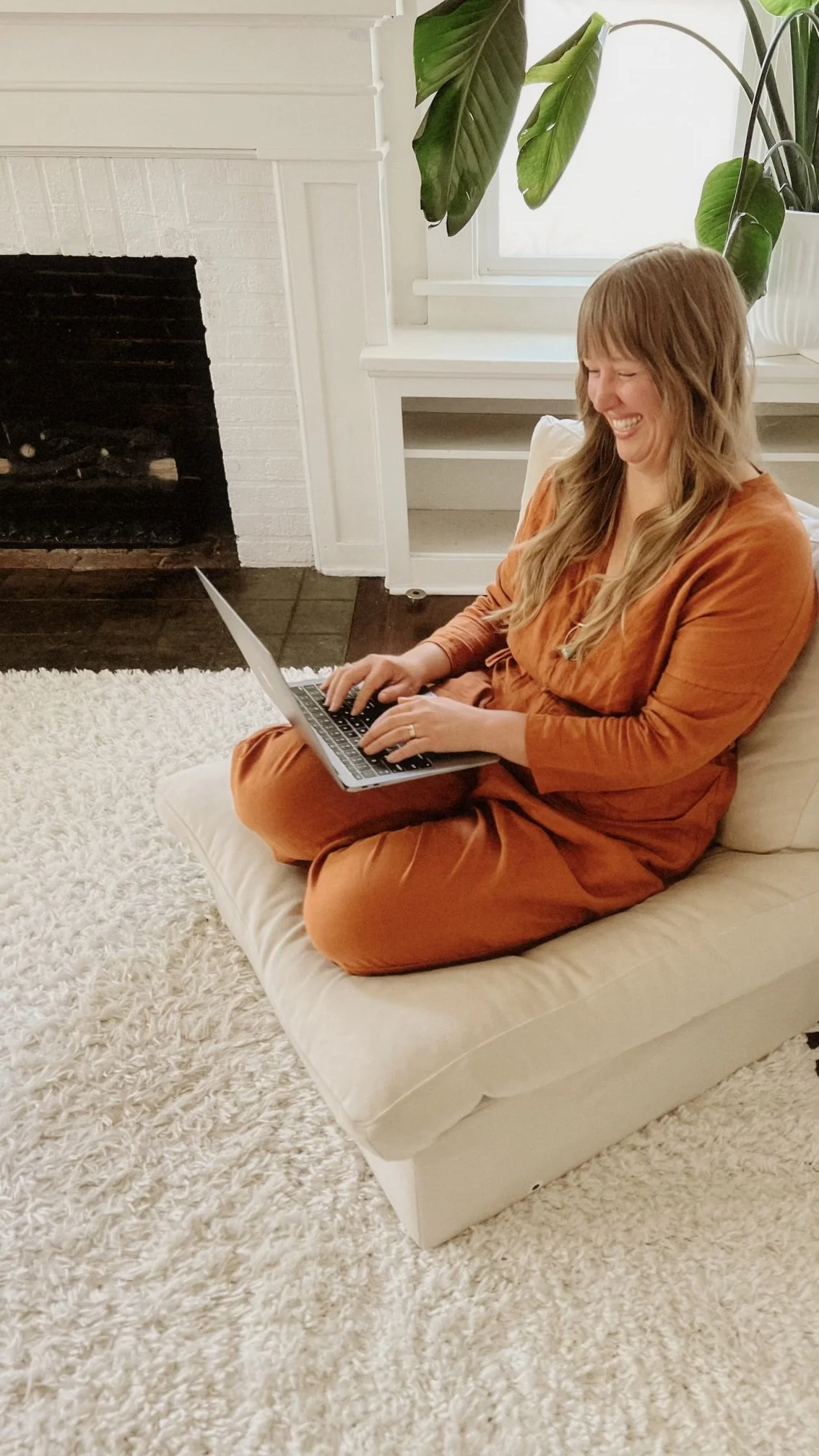 A woman with long, wavy blonde hair sitting on a beige couch, wearing an orange-brown dress, using a laptop while smiling and laughing in a cozy living room with a fireplace, white wall, and large green leafy plants.