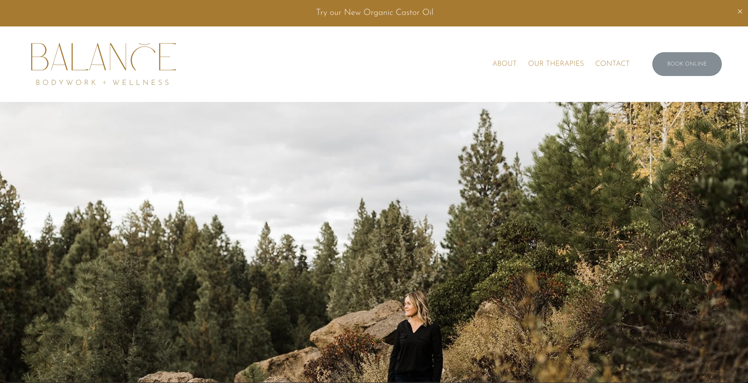 Woman standing outdoors among trees and rocks in a natural landscape with cloudy sky.