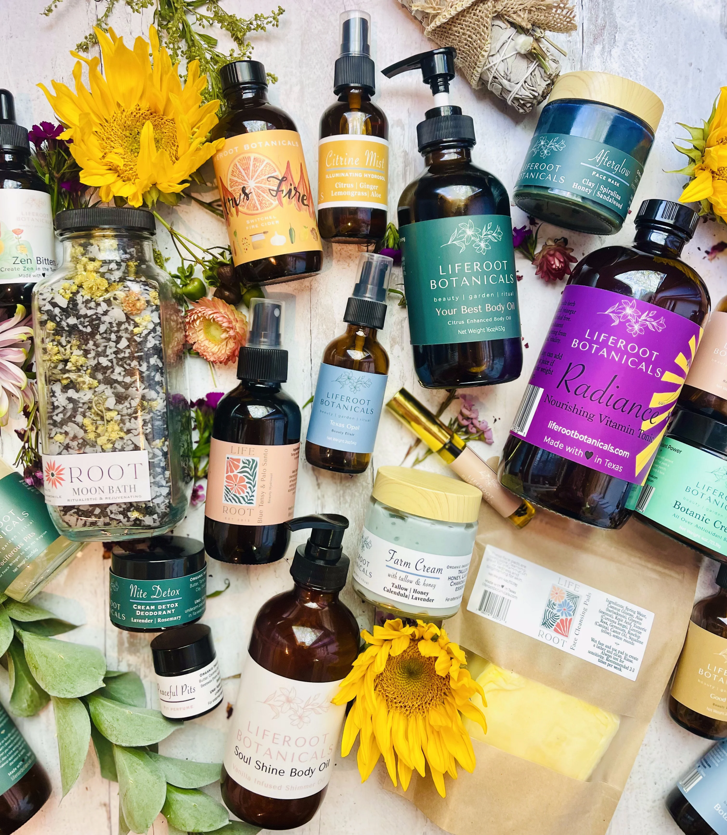 An assortment of natural skincare and wellness products, including bottles of body oils, sprays, and creams, arranged with yellow flowers, green leaves, and decorative packaging.