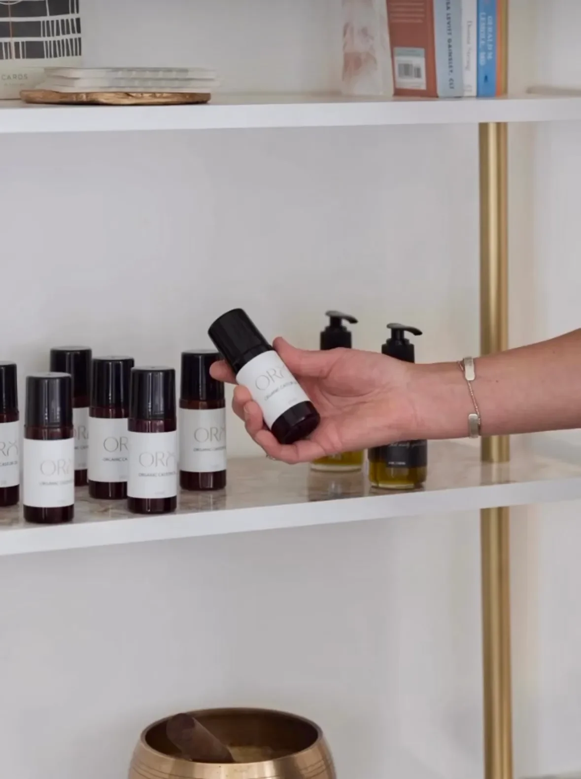 Hand holding a small dark bottle labeled 'OR' in front of a shelf with multiple similar bottles and some soap dispensers.