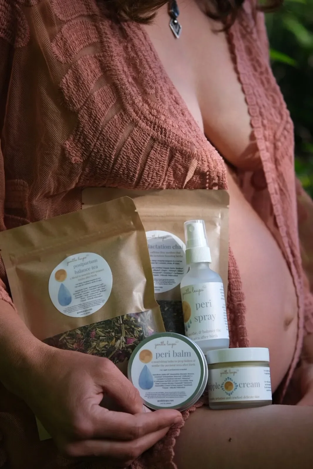Person in a pink lace top holding a collection of skincare and wellness products, including a Peri spray, Peri balm, a spray bottle, and herbal tea, with a blurred green background.