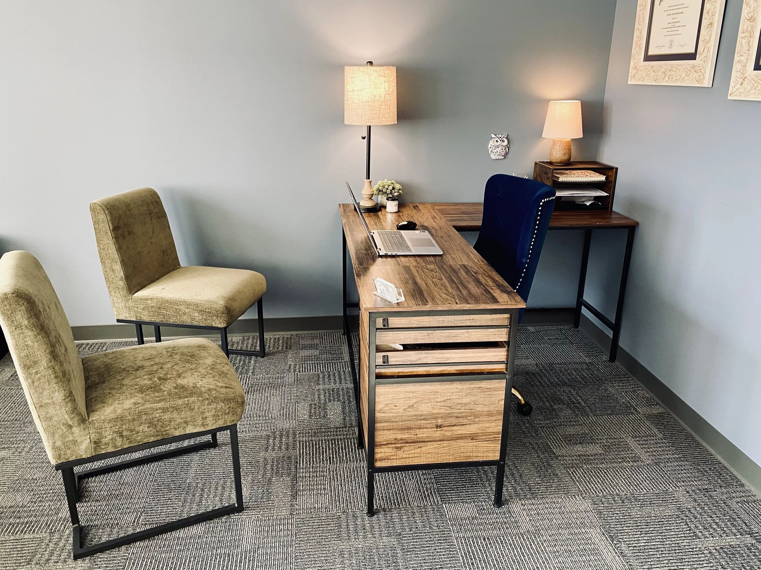 Home office with a wooden desk, two beige chairs, a black upholstered chair, two table lamps, and framed certificates on a blue wall.