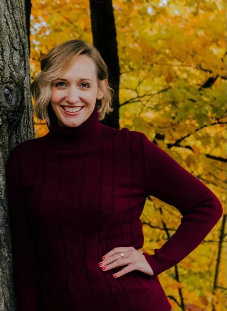 A woman with blonde, wavy hair smiling and wearing a maroon turtleneck sweater, standing outdoors in front of a tree with yellow and orange autumn leaves.