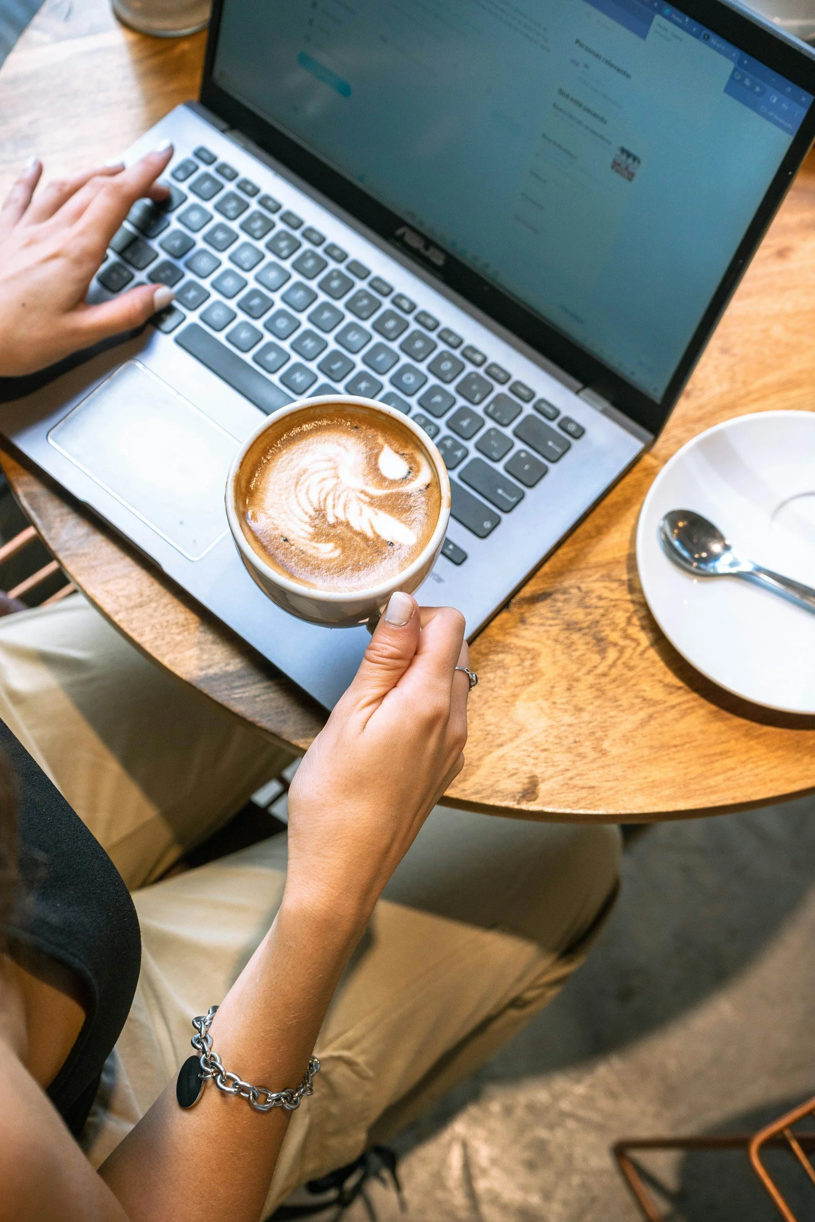 A person working on a laptop at a wooden table, holding a cup of coffee with latte art, and a white plate with a spoon nearby.