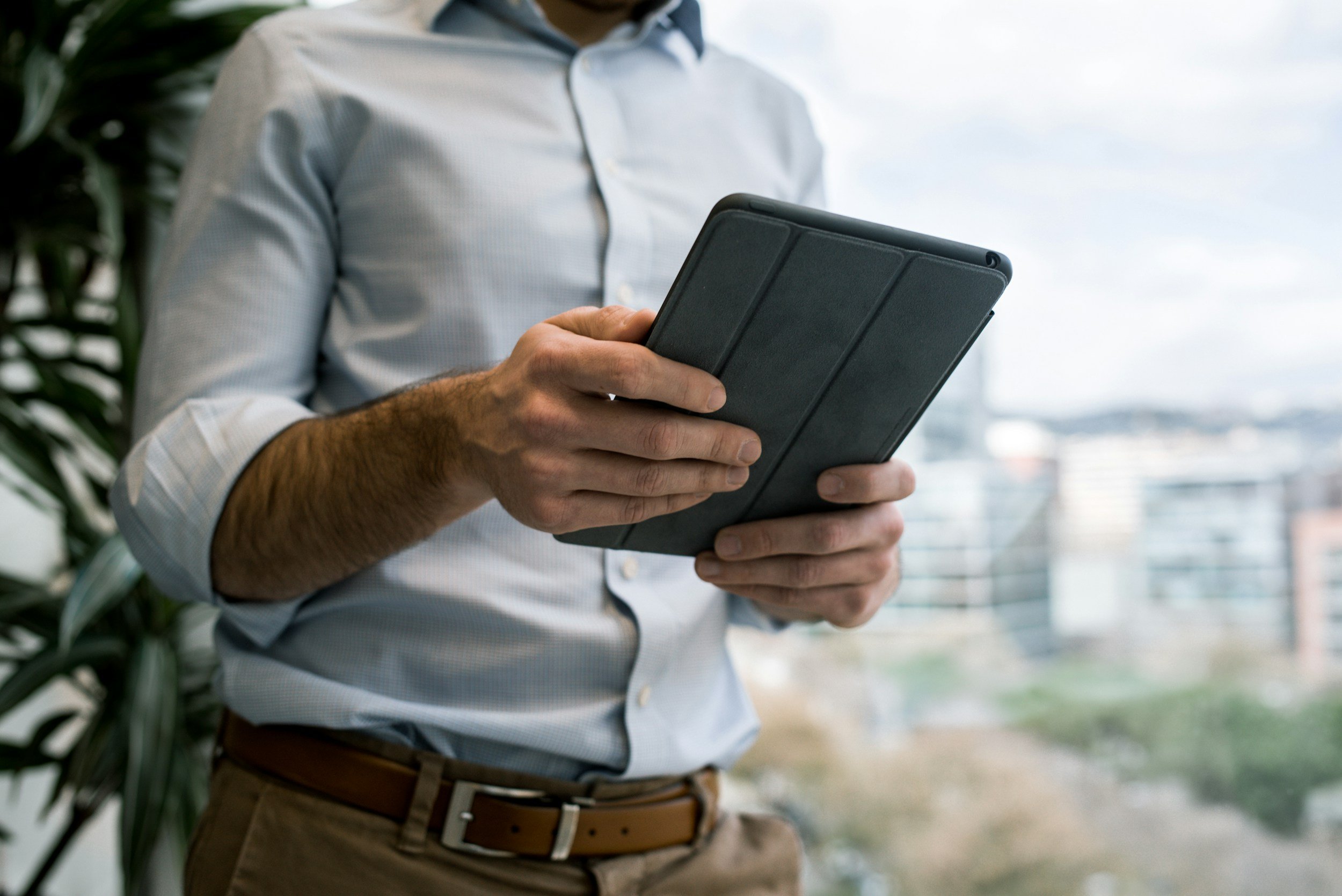 A man wearing a light gray button-up shirt and khaki pants holding a black tablet in front of a large window with a cityscape view.