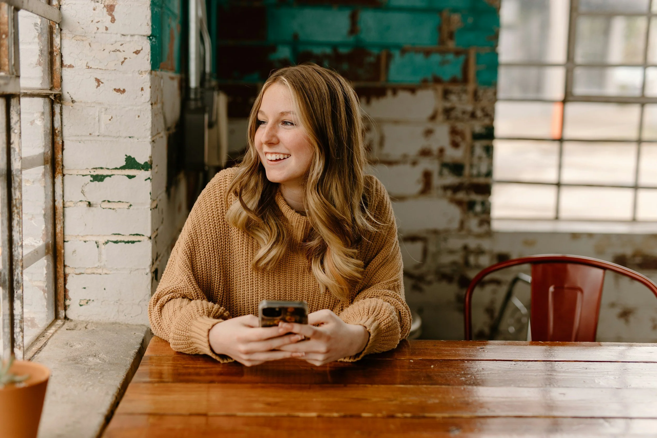A young woman with long wavy hair in a brown sweater sits at a wooden table in an industrial-style cafe, holding a smartphone and smiling while looking to her left.