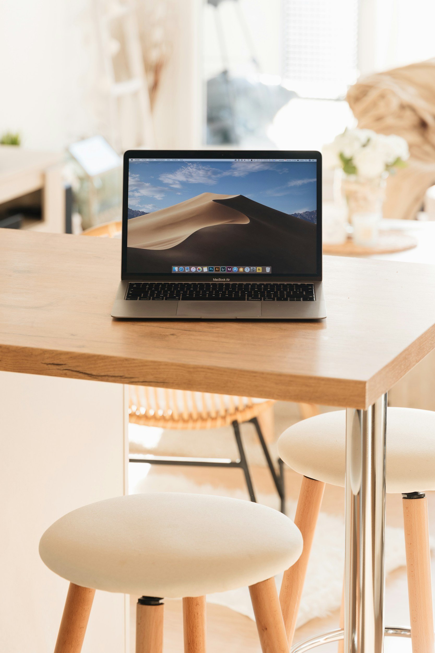 Open MacBook Air on a wooden table in a cozy, well-lit room with chairs and flowers in the background.