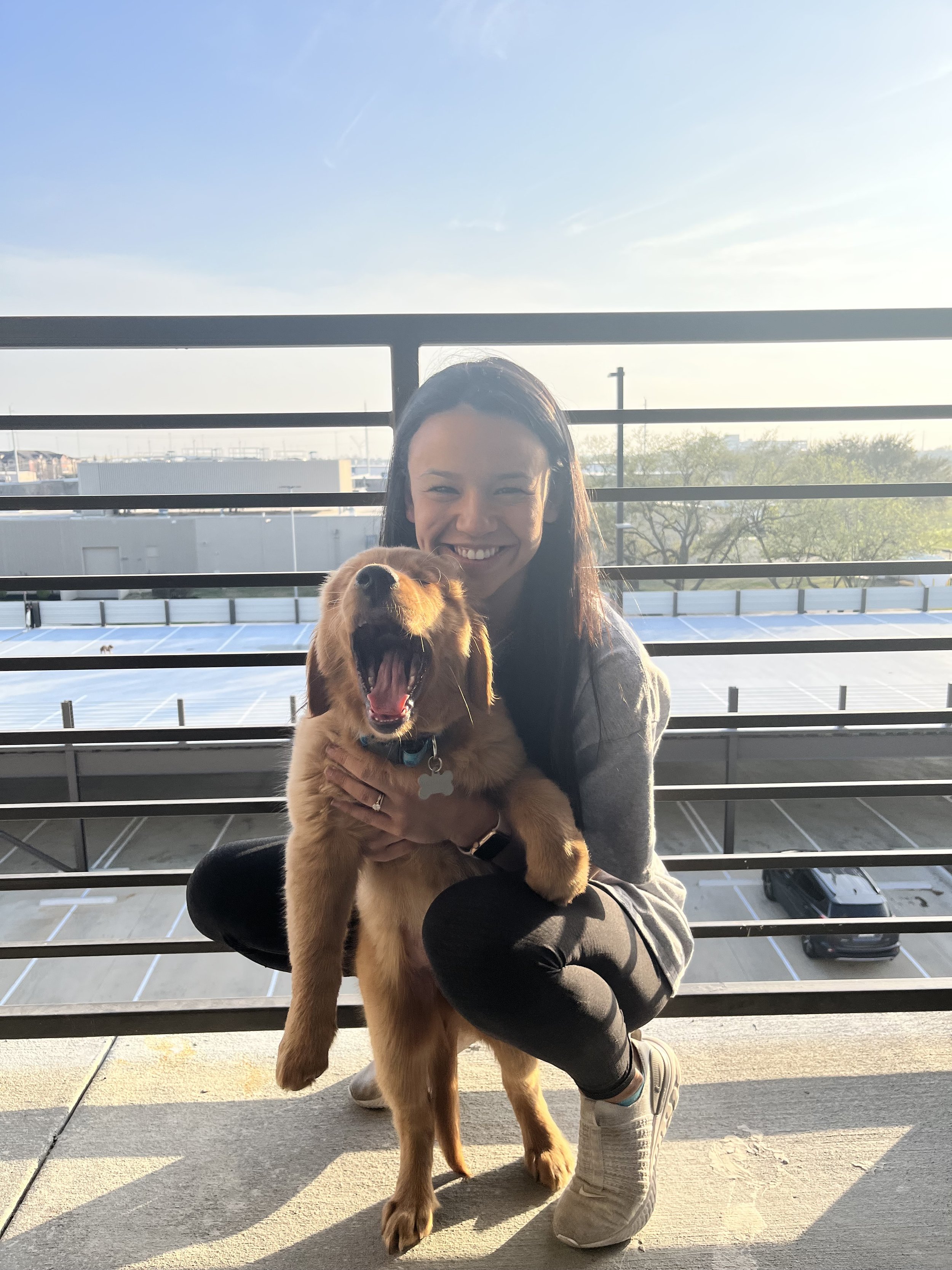 Woman smiling, holding a yawning golden retriever puppy on a balcony with a parking lot and clear sky in the background.