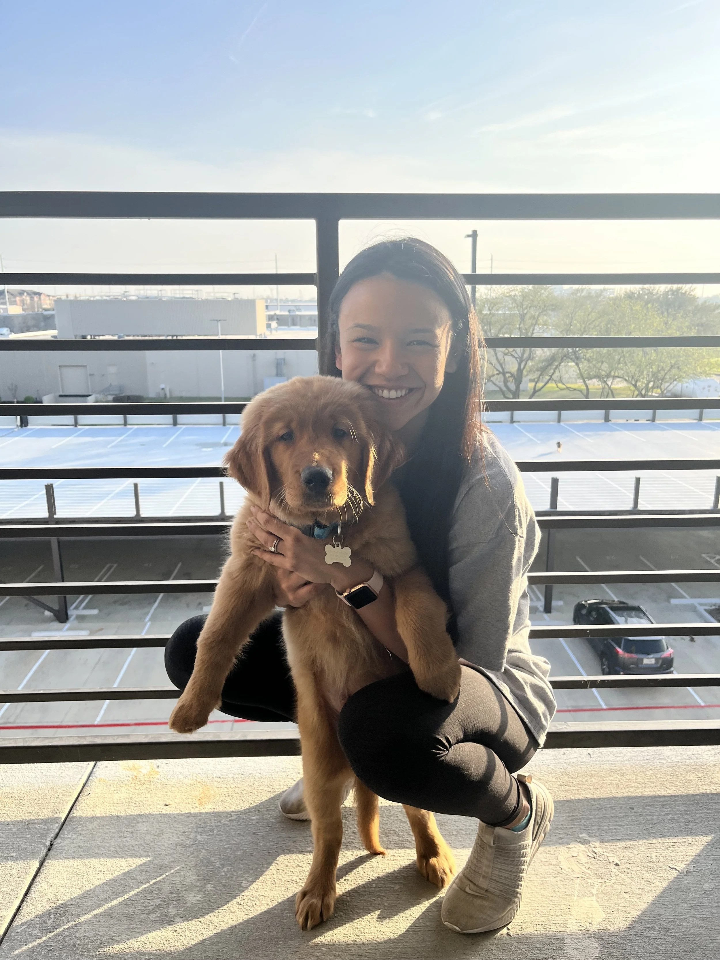 A woman crouching on a balcony holding a golden retriever puppy, with a parking lot and a building in the background during daylight.