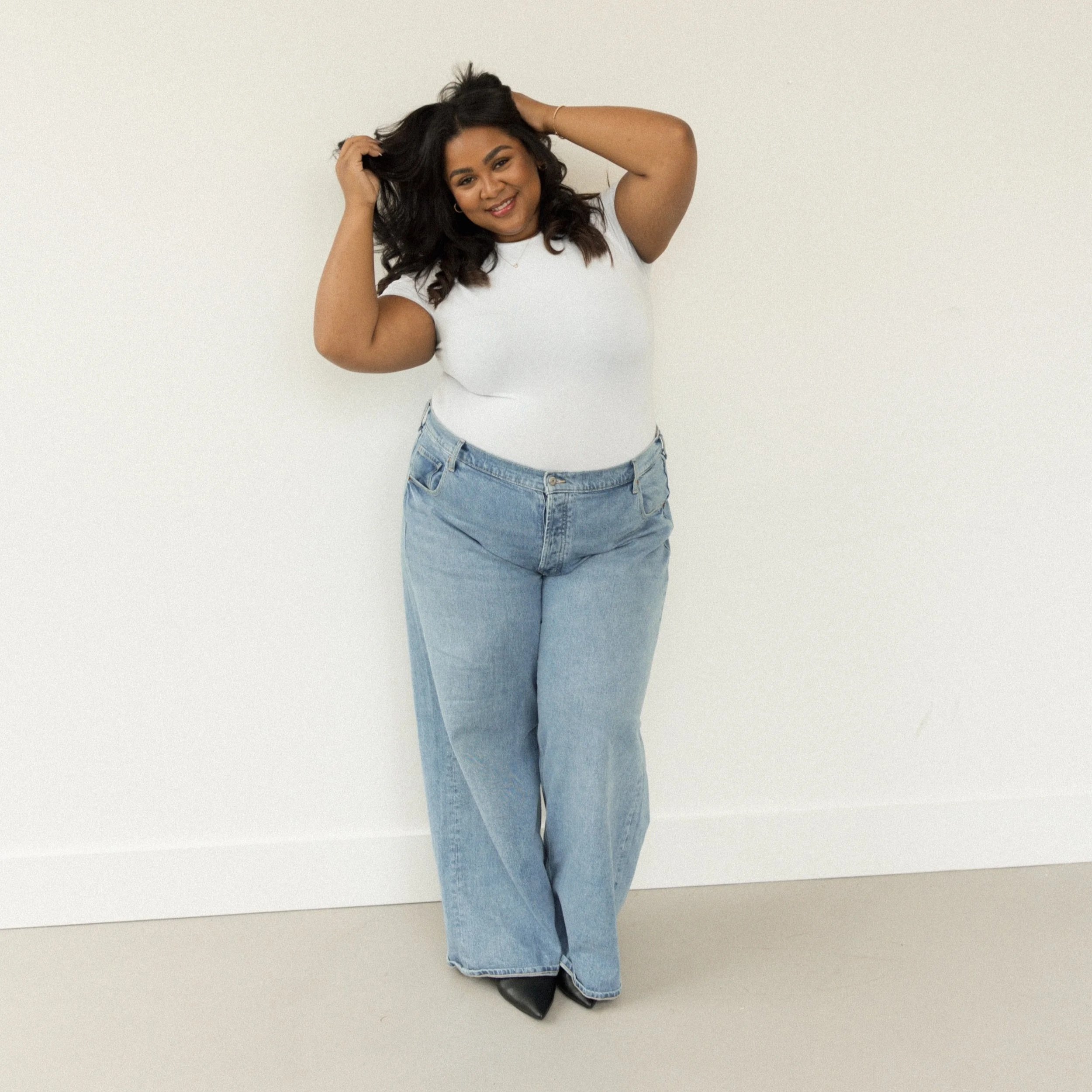 A woman with dark hair wearing a white t-shirt, light blue jeans, and black shoes poses against a white wall, smiling and touching her hair.