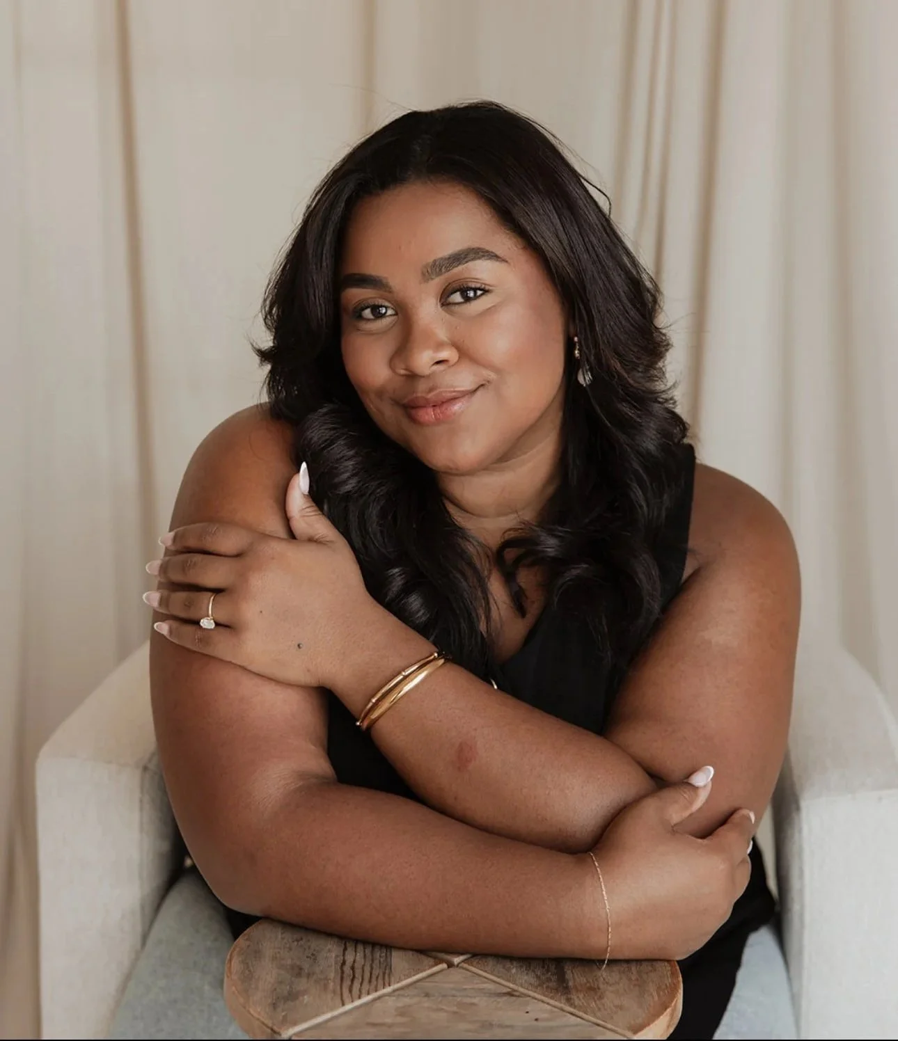 A woman with medium-length black hair smiling, sitting with her arms crossed over a wooden table, wearing a black sleeveless top and gold jewelry, in front of a beige curtain.