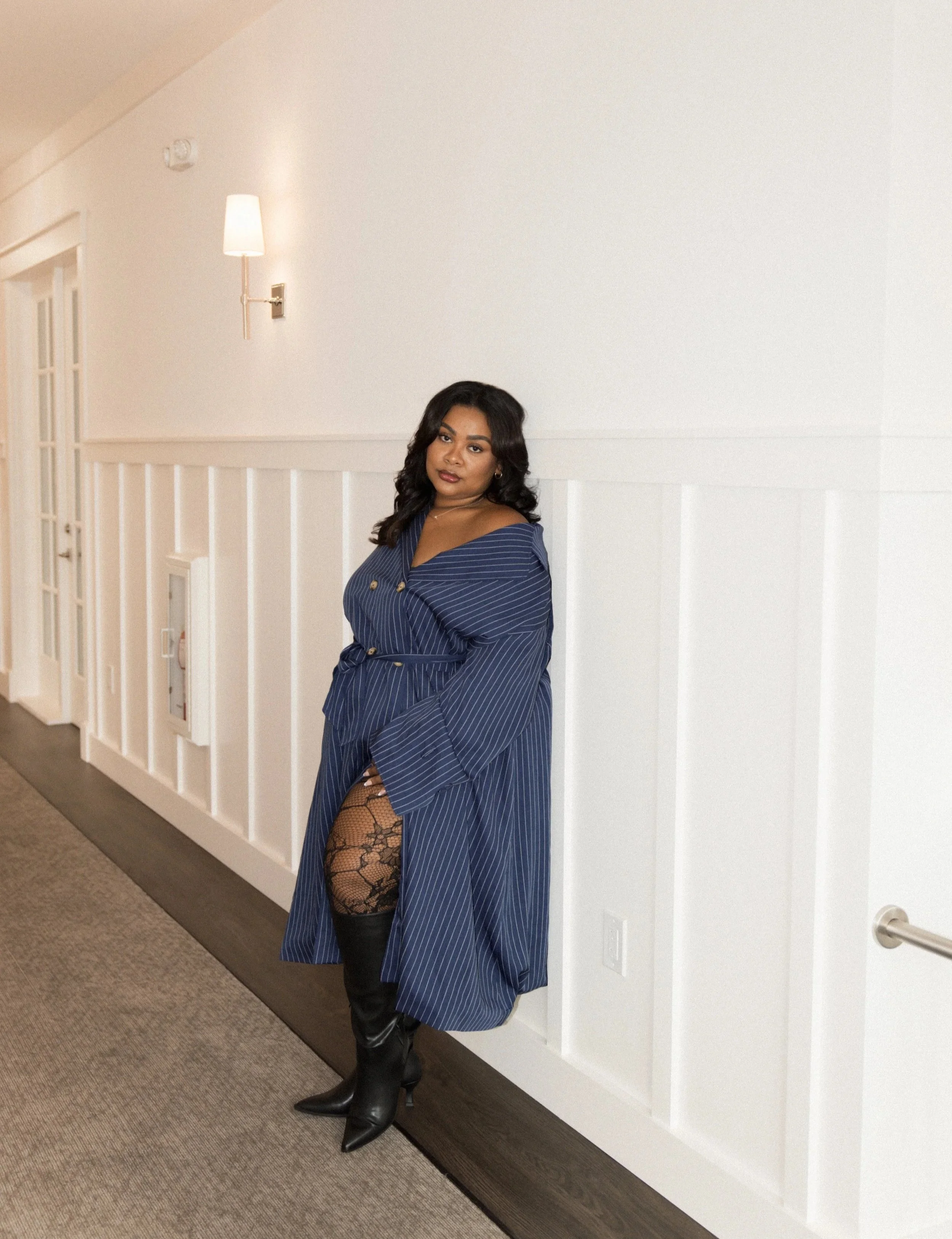 Woman in a blue pinstripe dress with black lace stockings and knee-high black boots standing against a white paneled wall in a hallway.