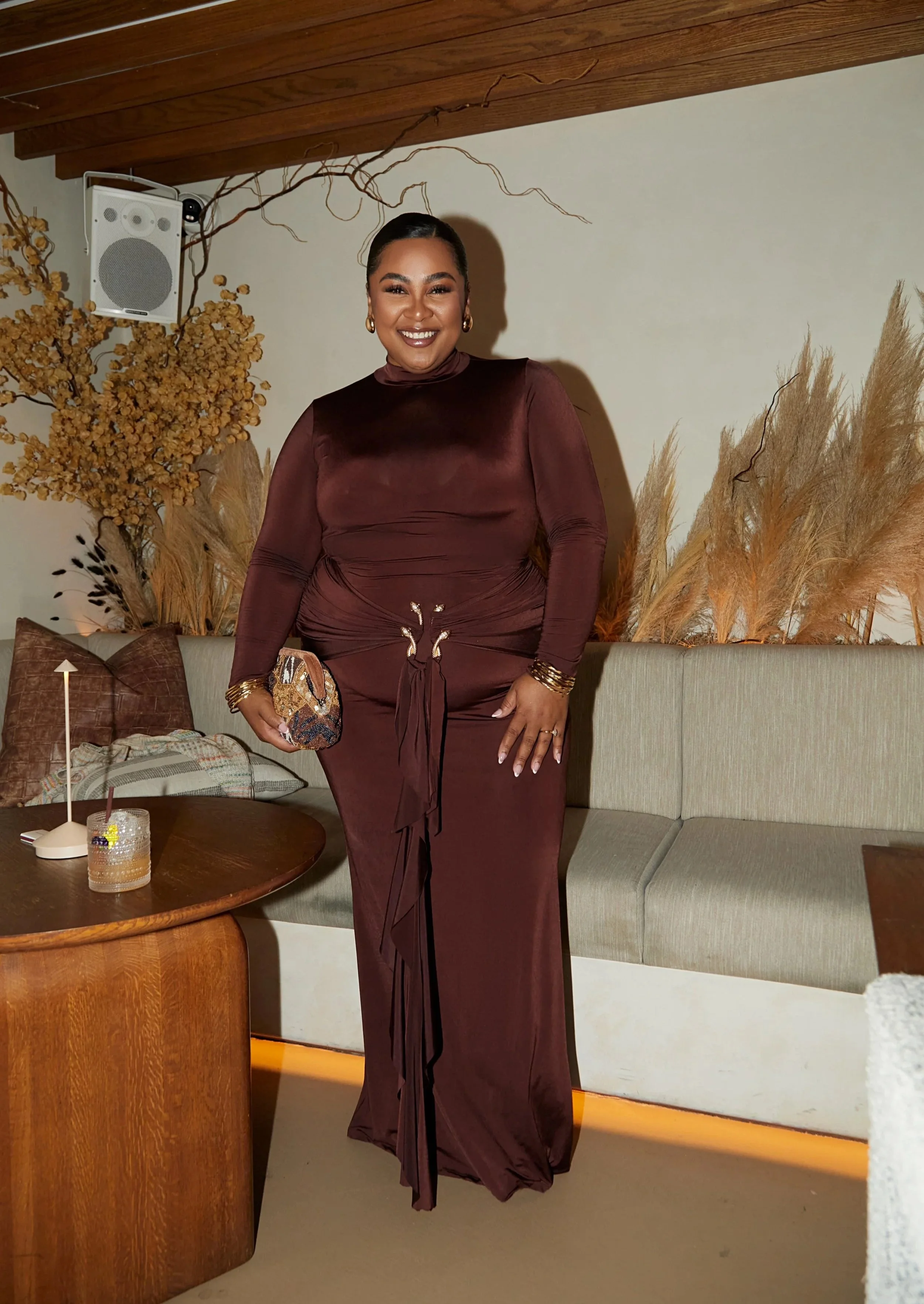 A woman wearing a long, dark brown dress with gathered details standing in a decorated indoor setting with dried plants and a beige sofa behind her. She is smiling and holding a clutch purse.