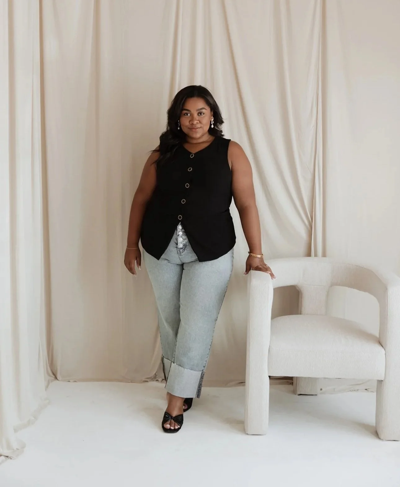 Woman in black sleeveless top and gray pants standing next to a cream-colored armchair in a room with cream curtains.