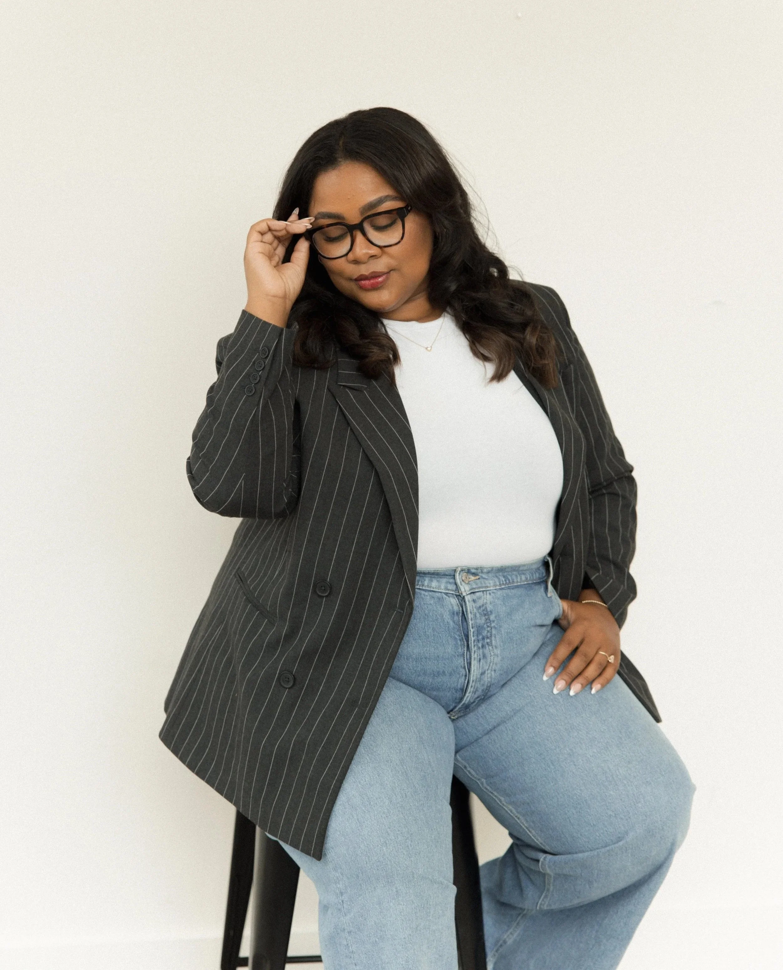 A woman with glasses, wearing a black pinstripe blazer, white top, and light blue jeans, sitting on a black chair against a plain white wall, looking down and adjusting her glasses.