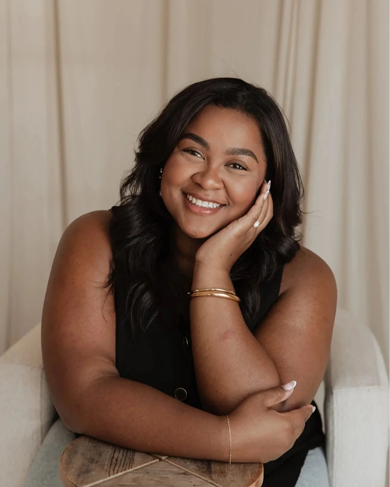 A young woman with dark hair smiling, resting her face on her hand, wearing a black sleeveless top, gold bracelets, and a ring, seated in front of beige curtains.