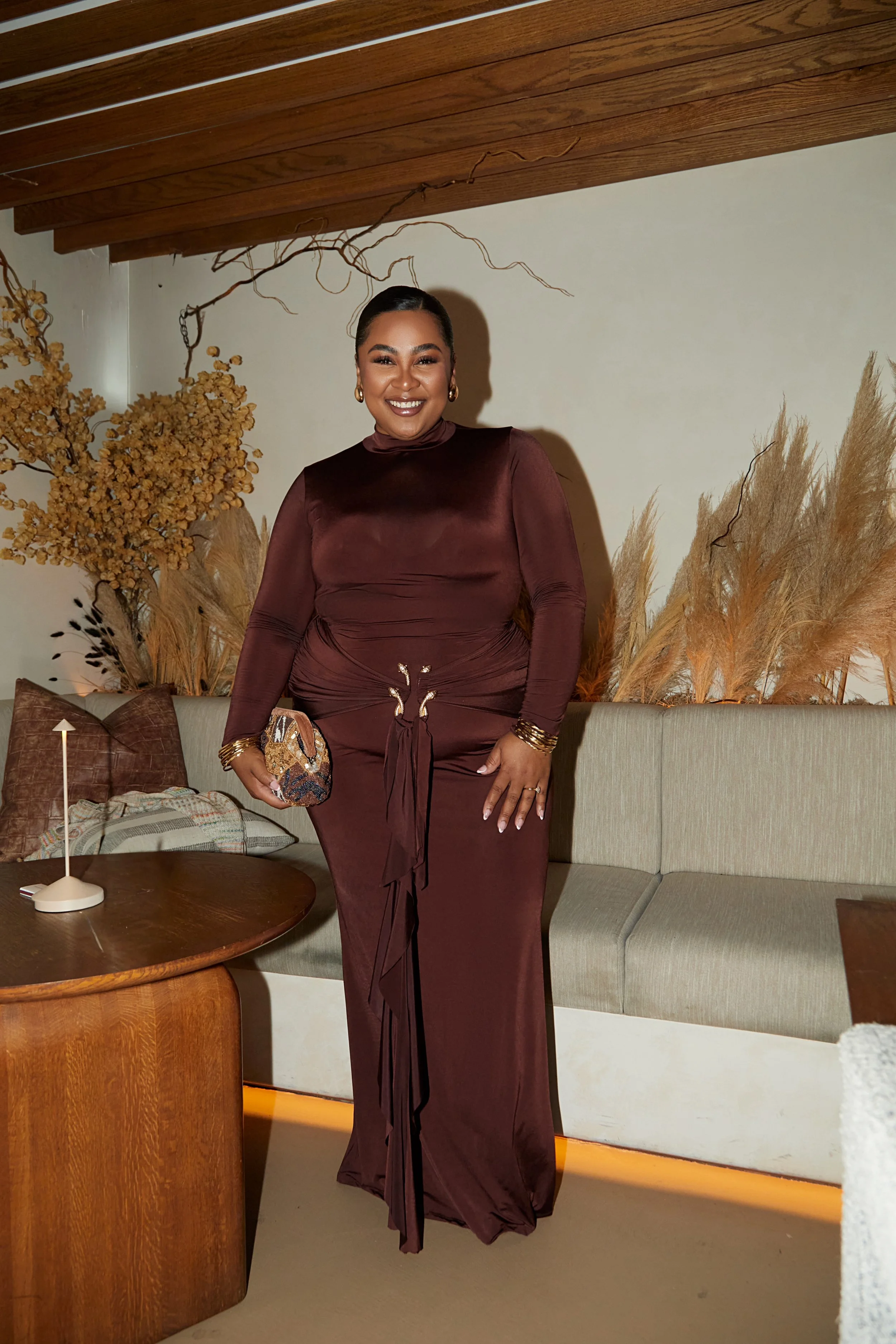 Woman in a long, brown satin dress standing in a cozy, decorated room with dried botanical arrangements.