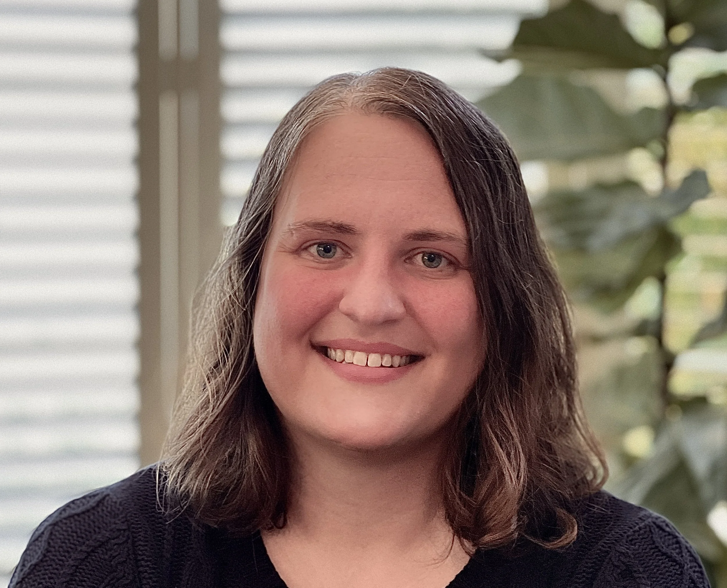 Portrait of a smiling woman with shoulder-length wavy hair, wearing a blue sweater, standing indoors near a window with blinds and some green plants in the background.