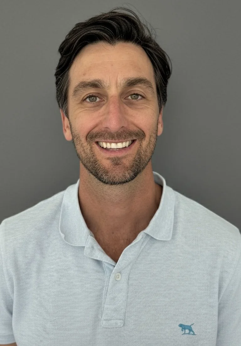 A smiling man with dark hair and a beard, wearing a light-colored polo shirt with a small blue logo of a dog on the left side, standing against a plain gray background.