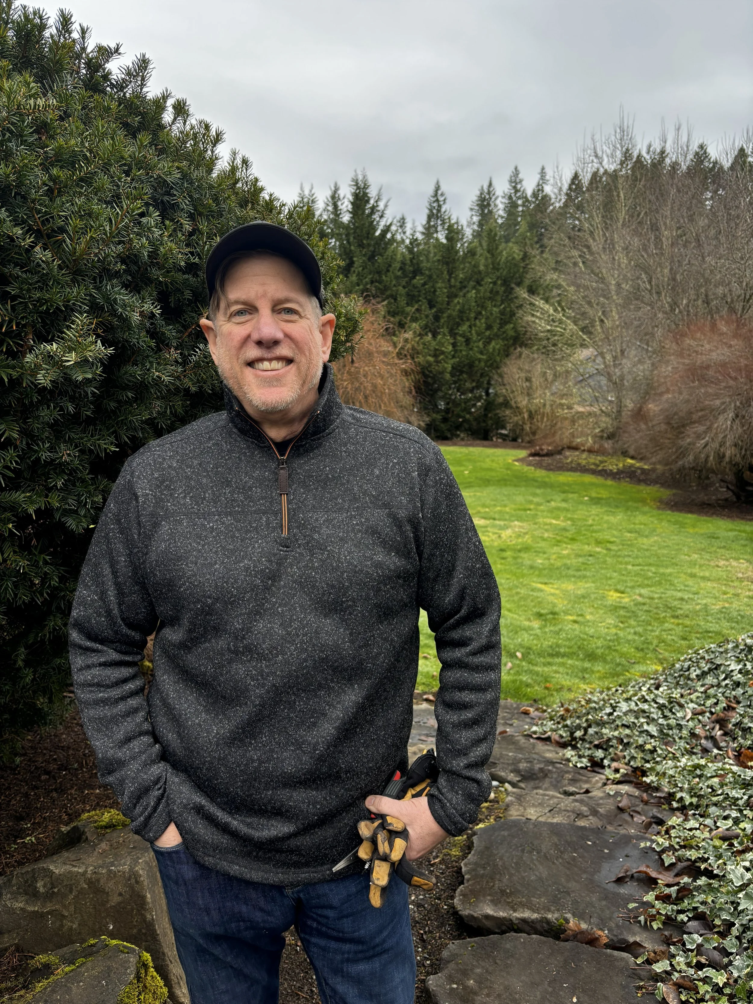 A smiling man outside in a garden with green grass, trees, and shrubs. He is wearing a black cap, dark gray sweater, and jeans, holding gardening gloves and tools.